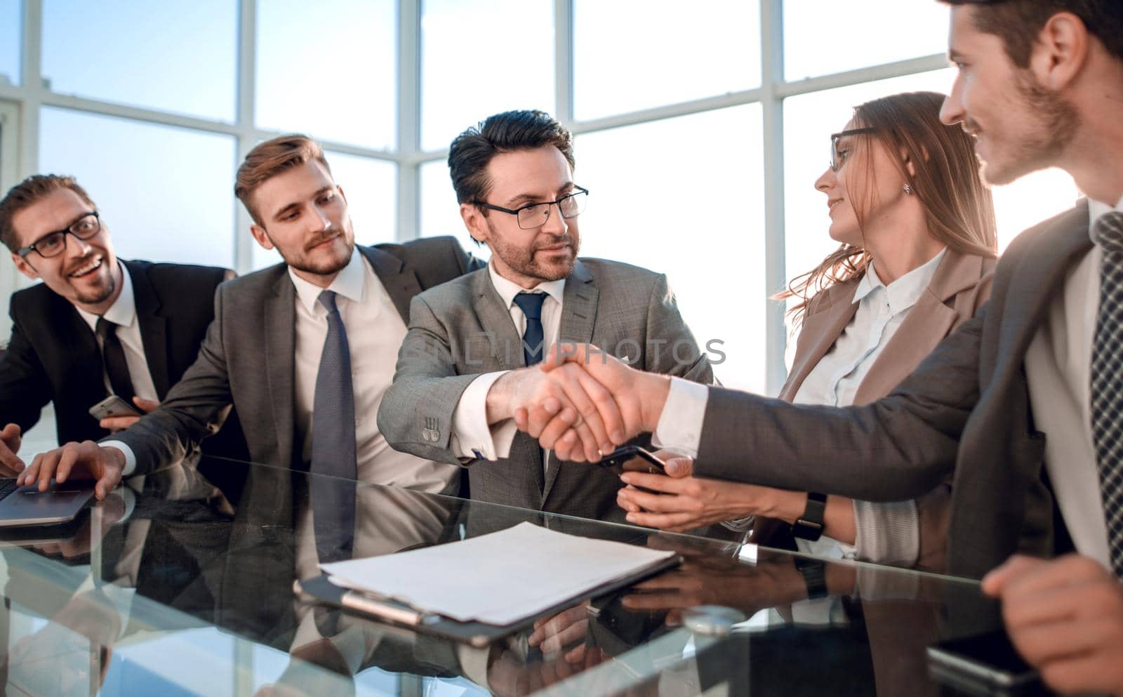Two confident young men shaking hands and smiling while sitting at the desk in office with two people working in the background