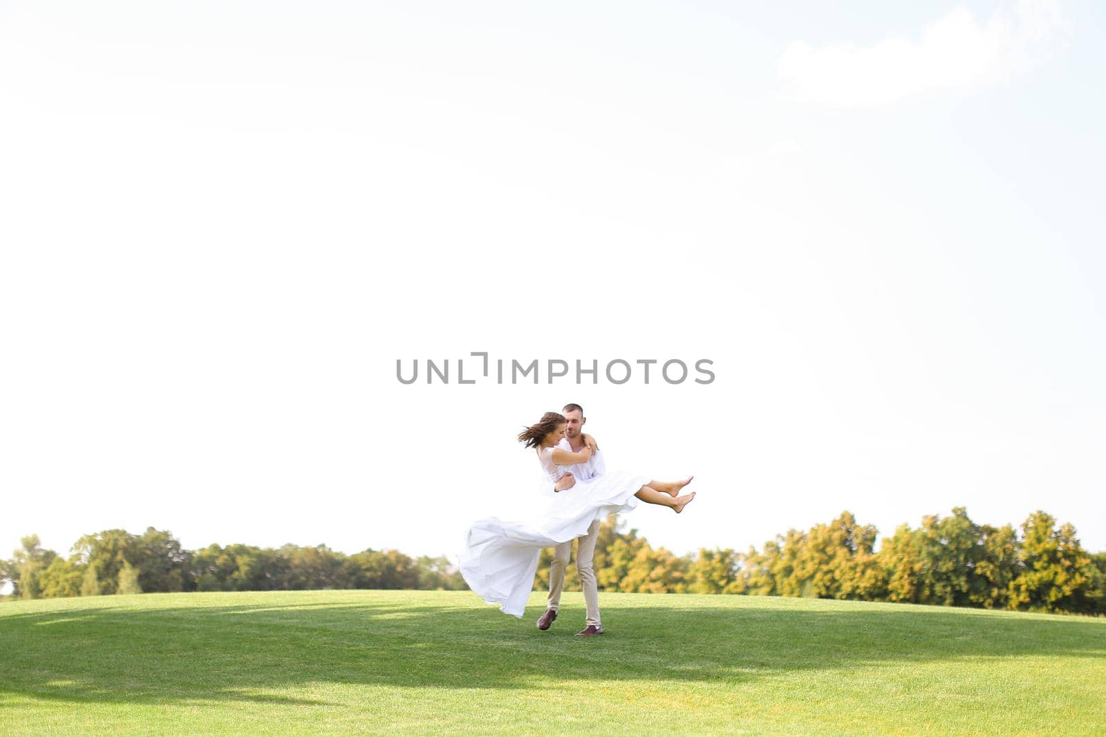 Happy groom holding bride on grass in white sky background. Concept of wedding photo session on nature.