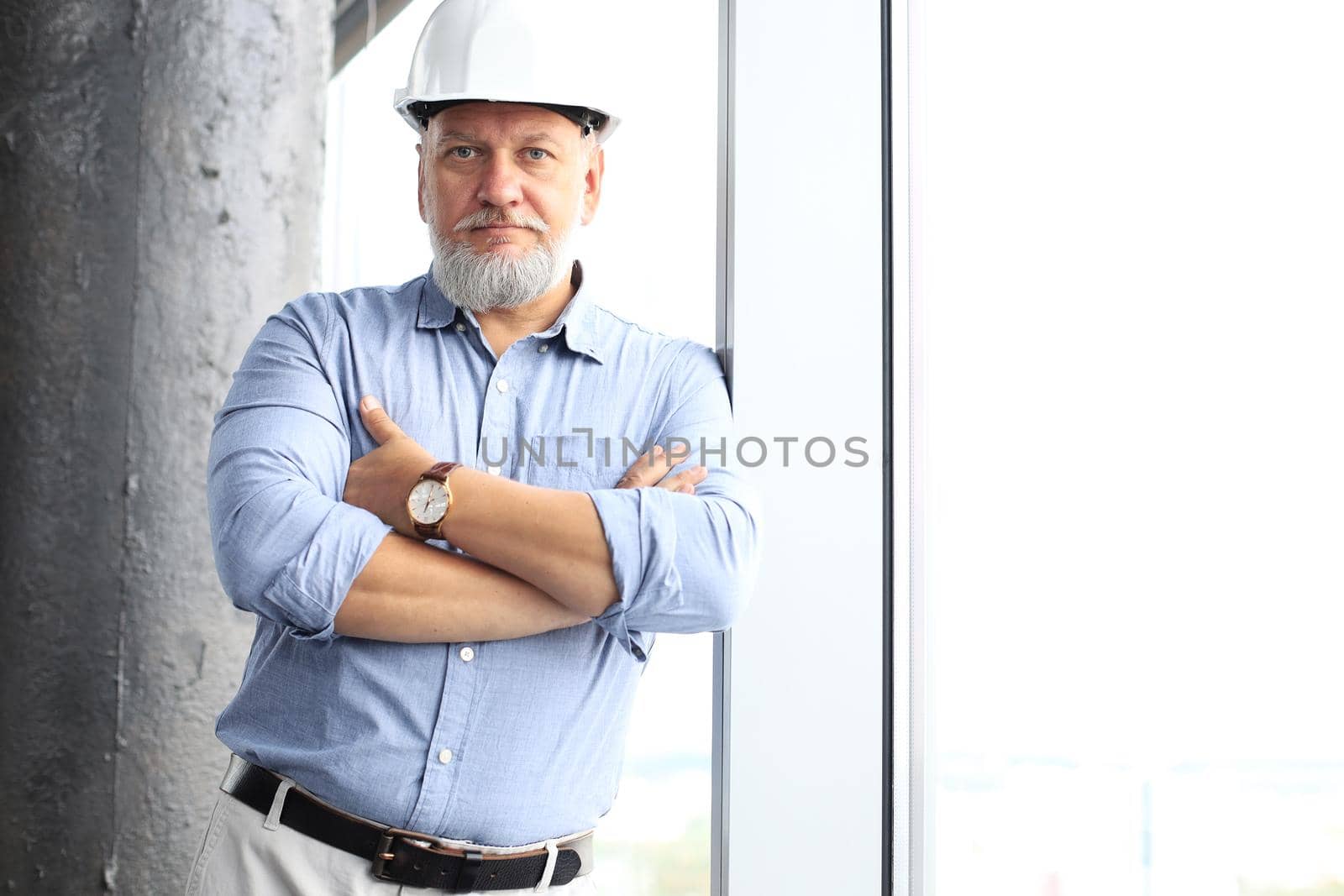 Thoughtful mature architect wearing hardhat and looking away while standing near the window in new modern building