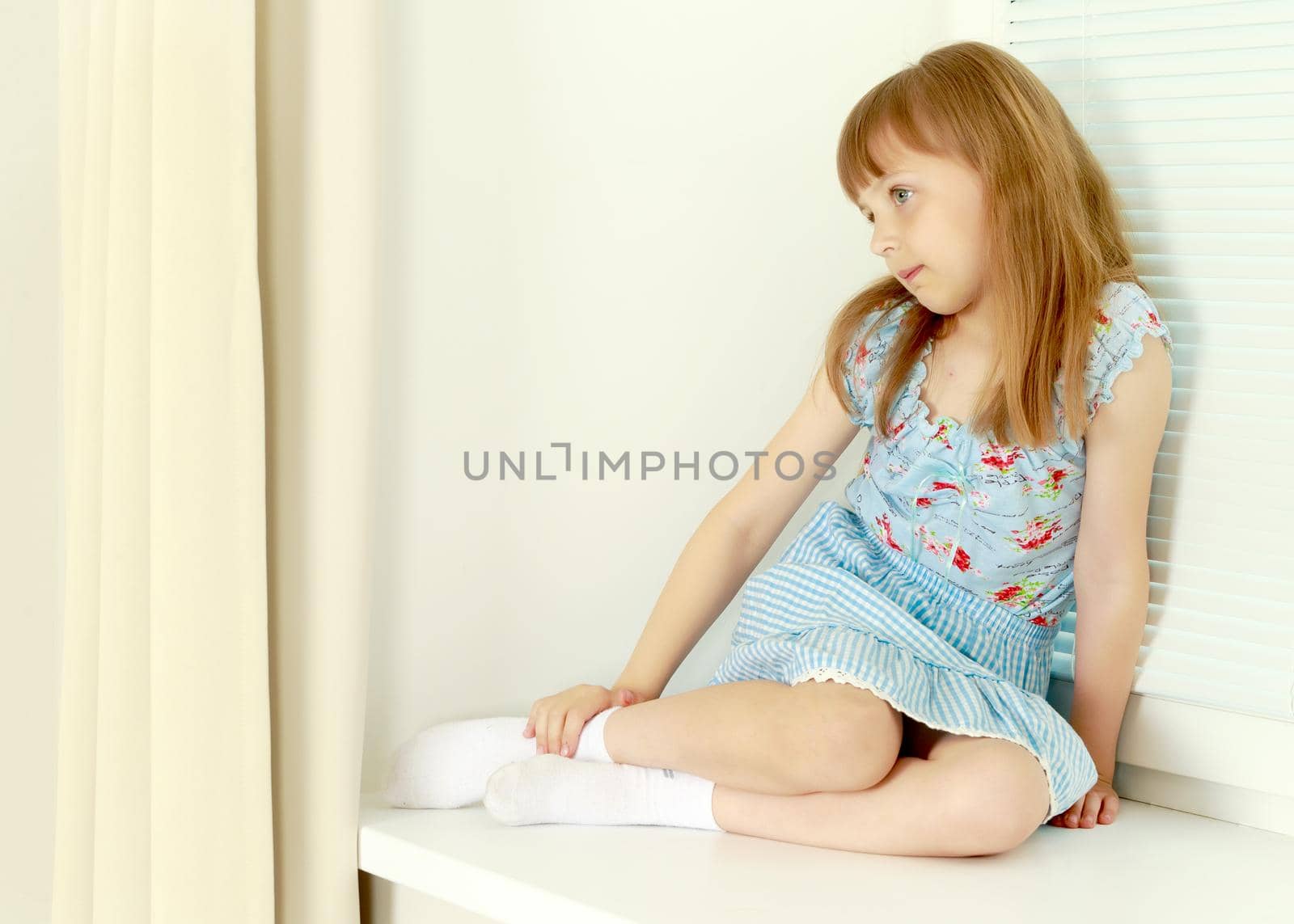 A nice little girl is sitting by the window with blinds. The concept of a happy childhood, construction, production of horizontal blinds.