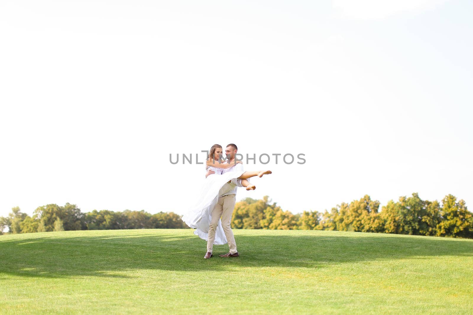 Caucasian groom holding bride on grass in white sky background. Concept of wedding photo session on nature.