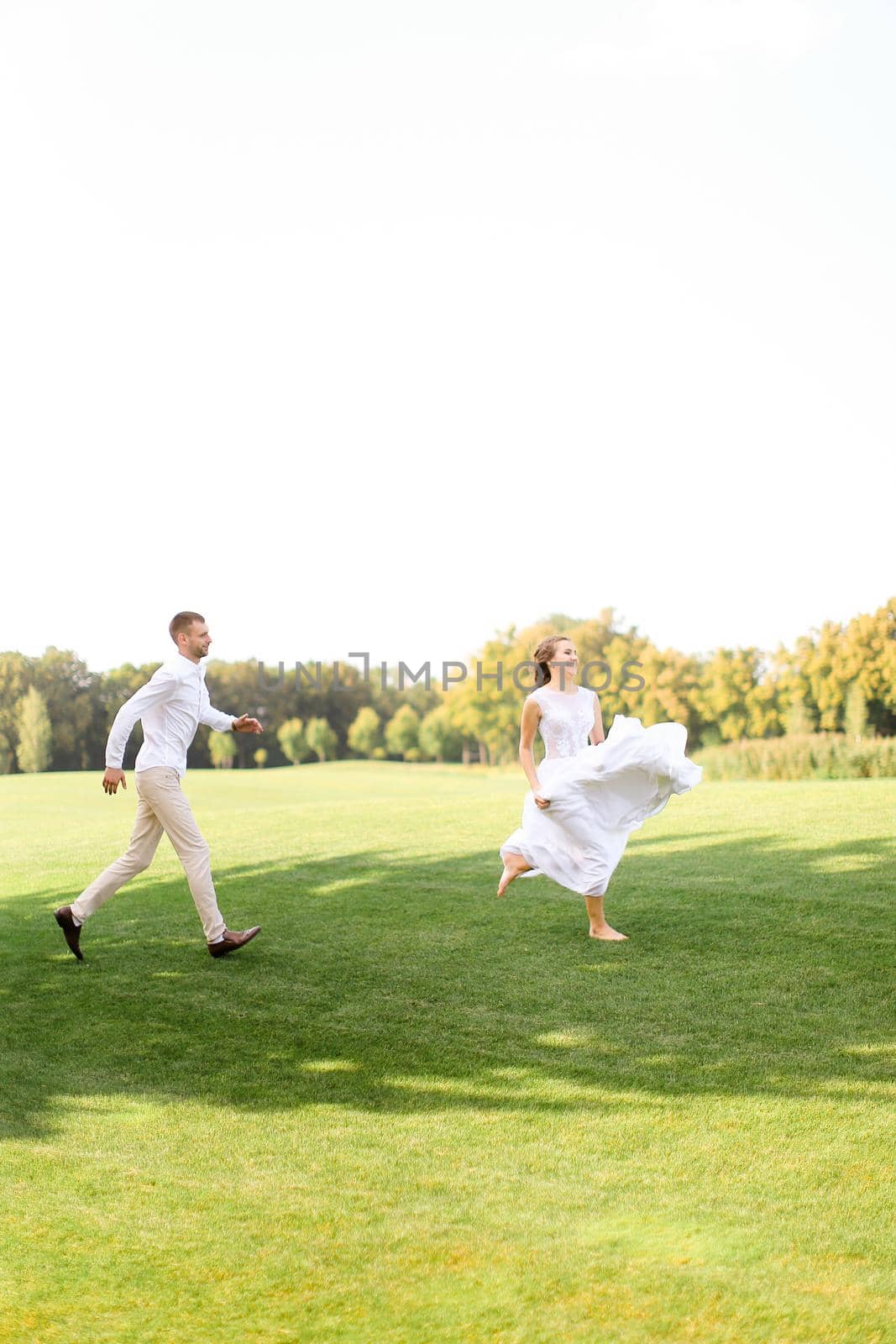 Groom and bride in white dress running and playing on grass. Concept of wedding photo session on open air and nature.