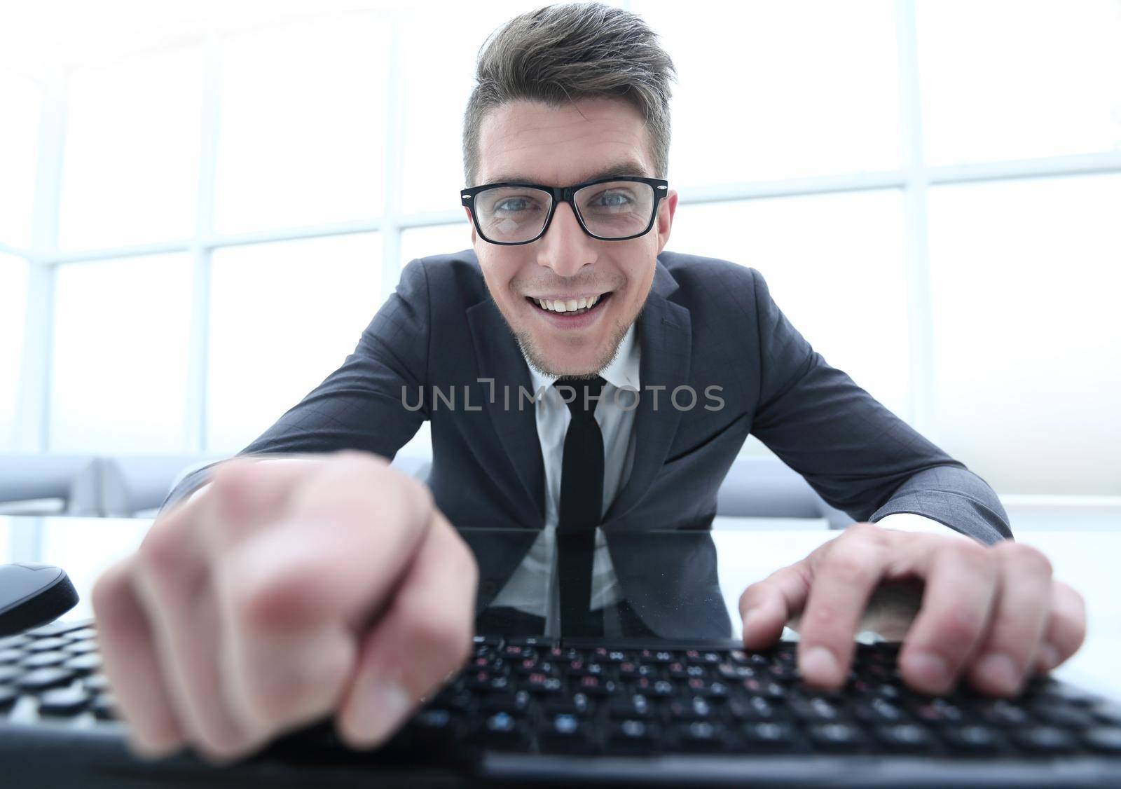 A mature man with gray hair wearing glasses siting at the table working with his laptop typing some documents looking with astonishment into camera pointing at you with his finger.