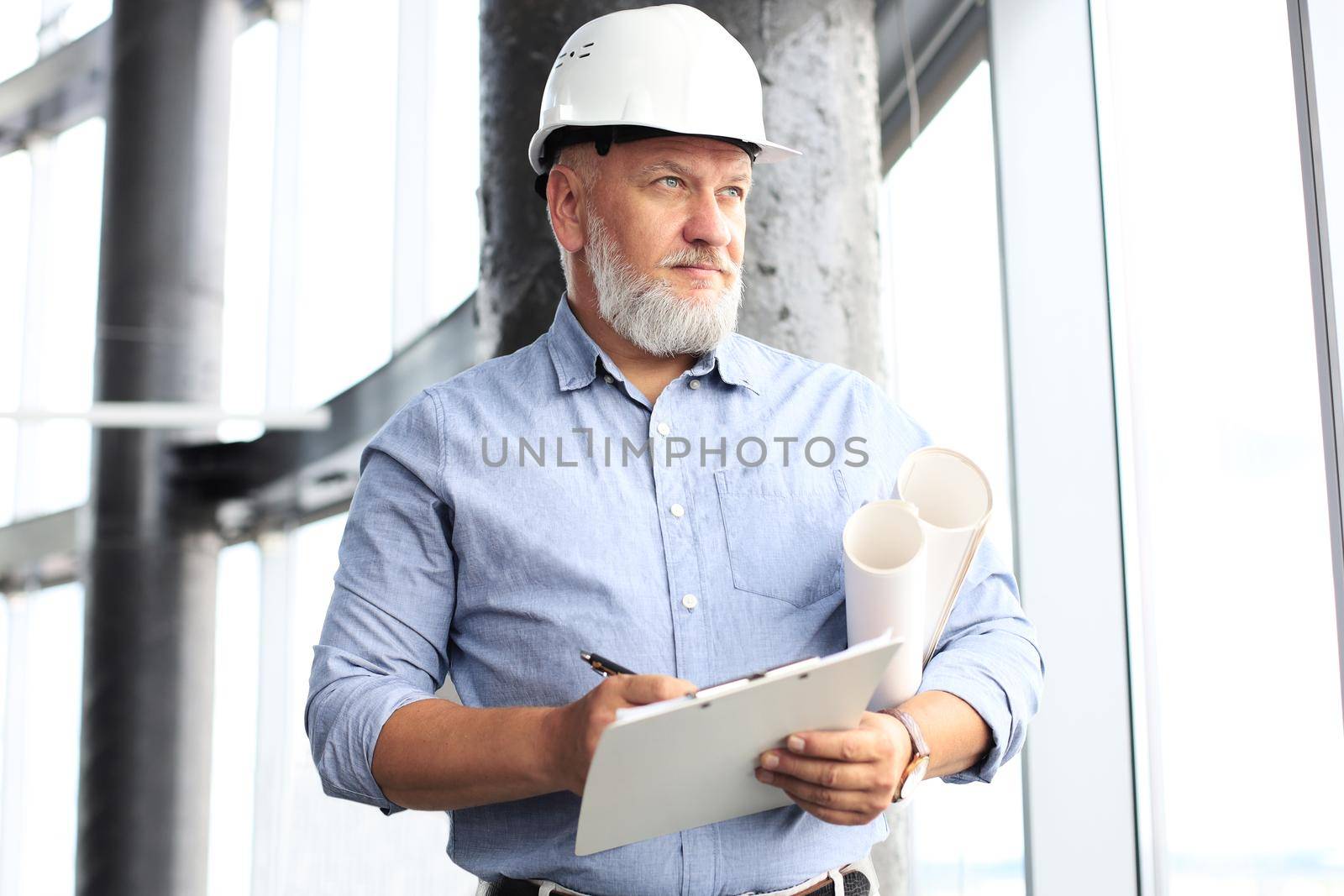 Confident mature business man in hardhat holding blueprint and looking away while standing indoors