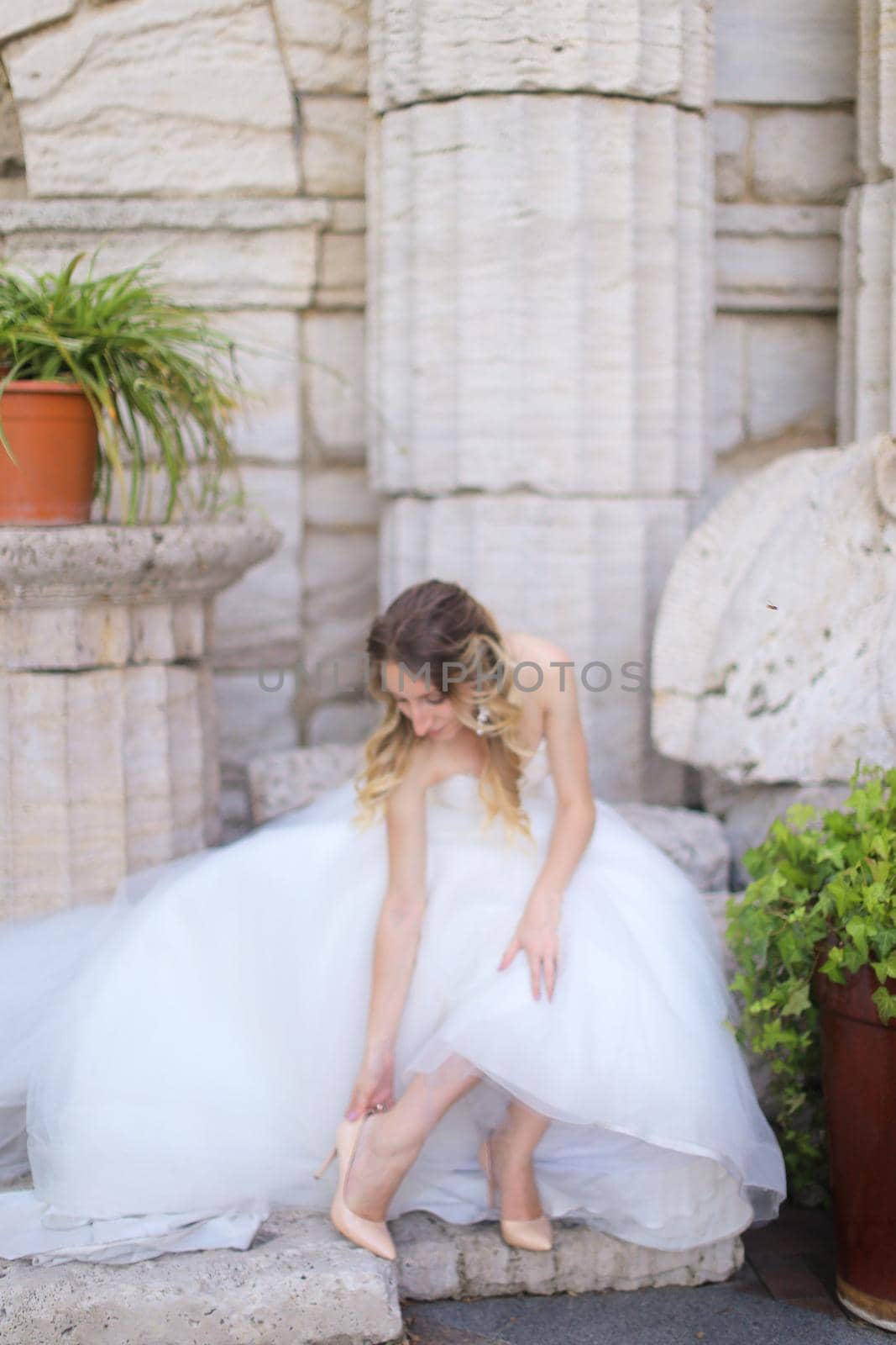 Caucasian nice fiancee sitting near ancient columns and and putting on shoes. Concept of bridal photo session and wedding.