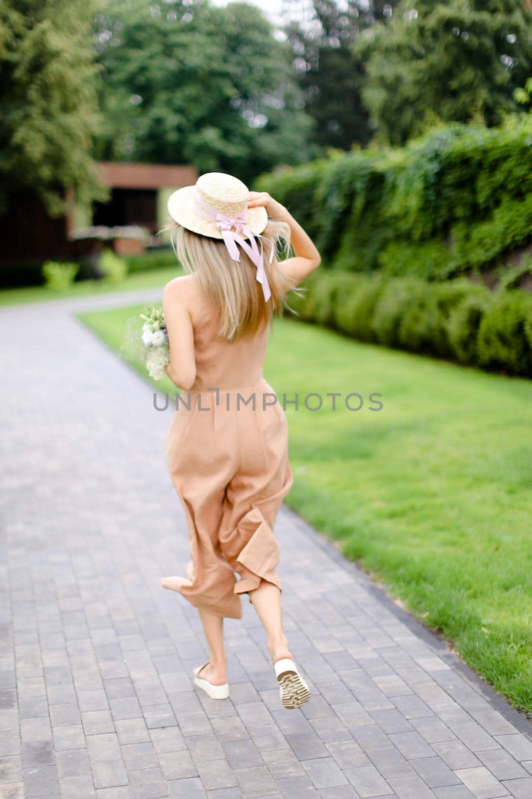 Back view of young blonde female person in body color overalls and hat standing in yeard with flowers. Concept of fashion and summer season.