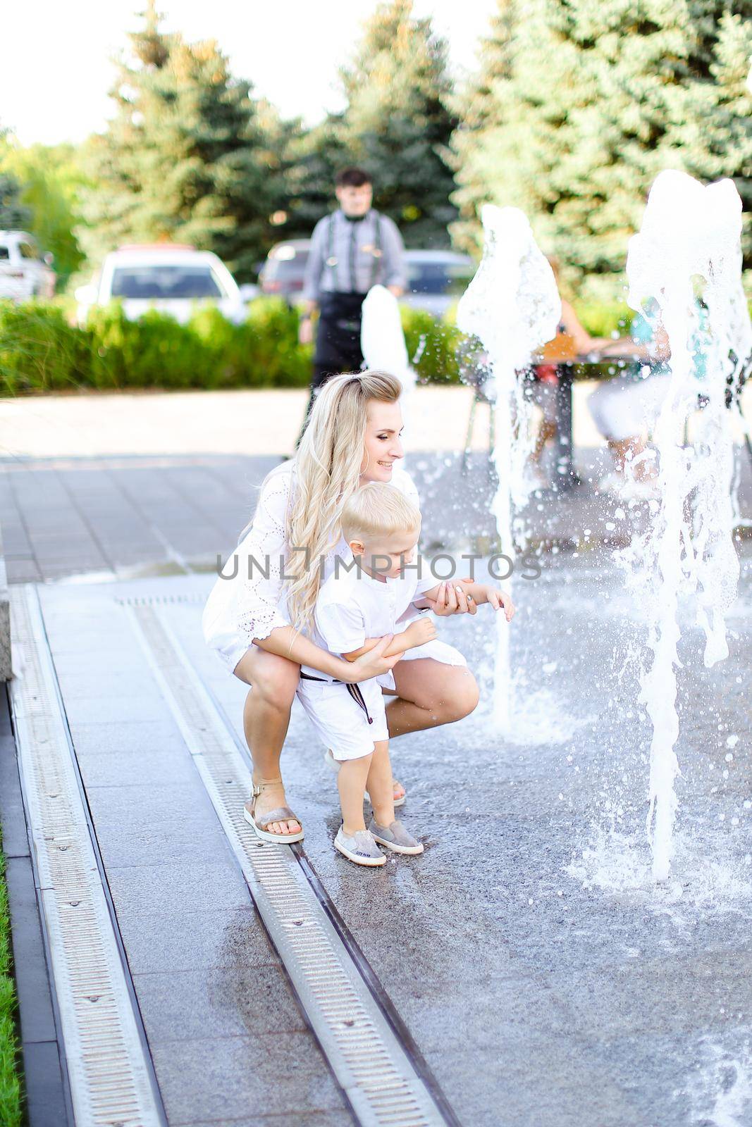 Young happy mother sitting with little baby outside near fountain. Concept of walking in city and motherhood.