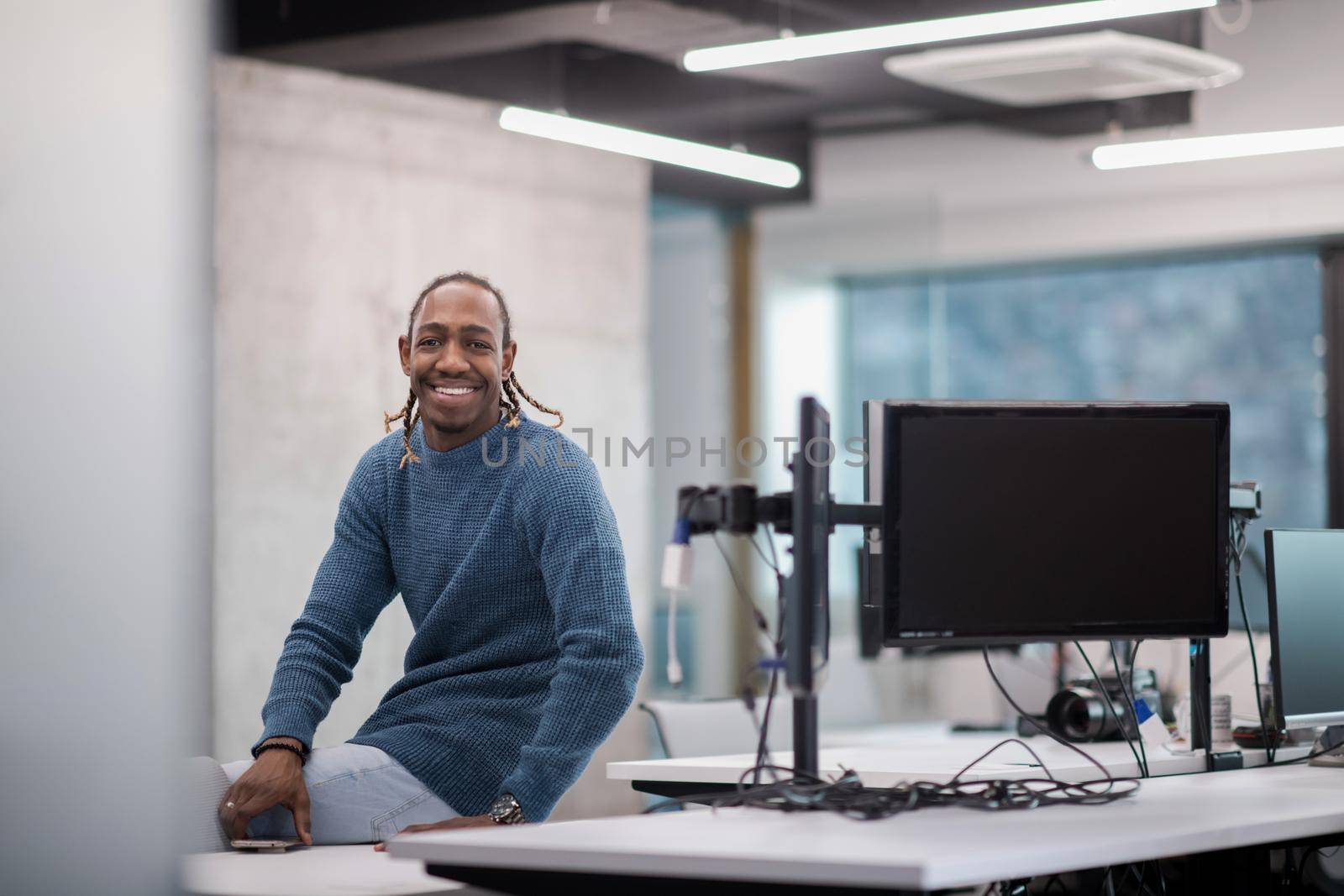 Portrait of young smiling african american male software developer sitting on office desk at modern startup office