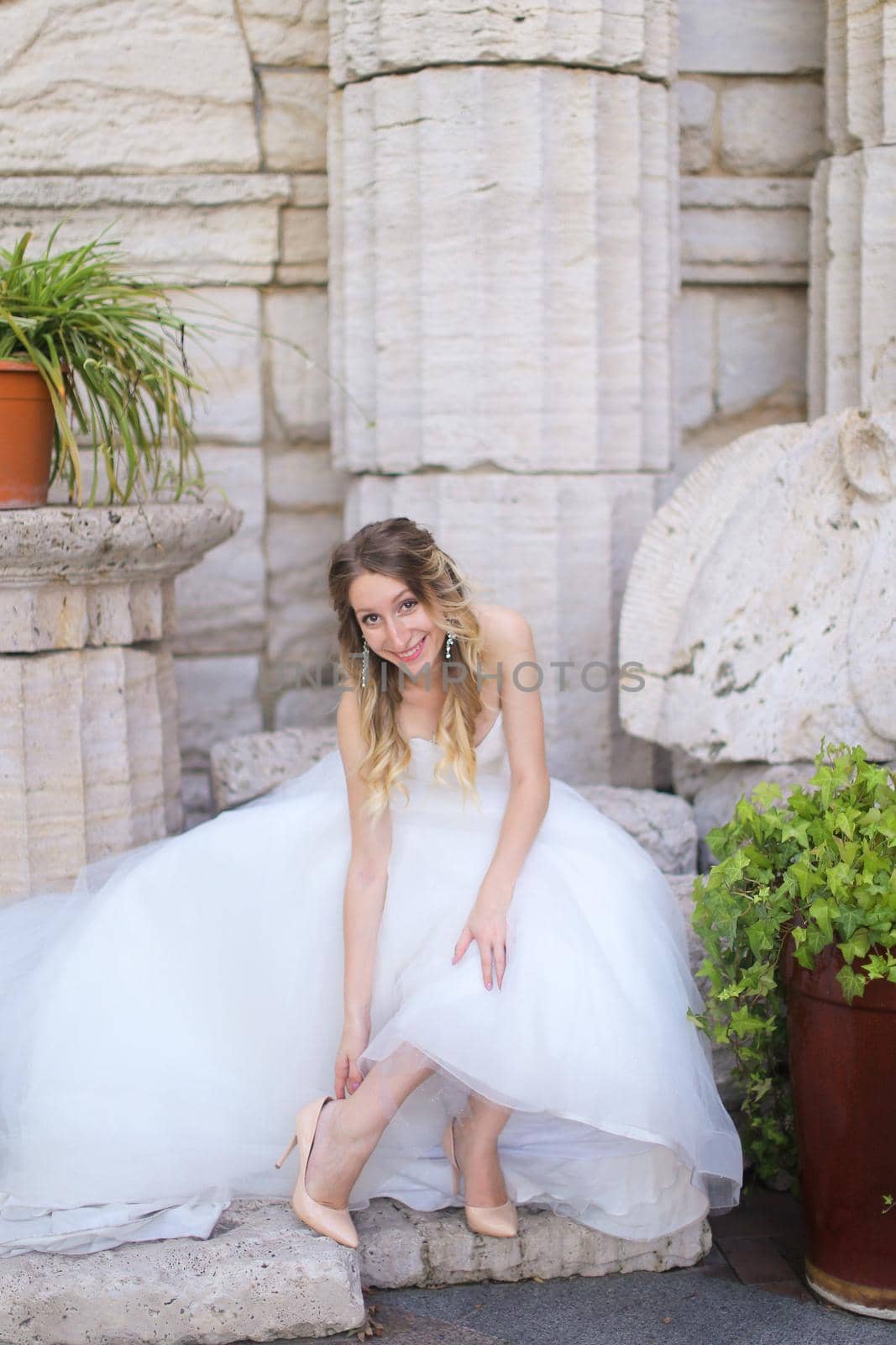 Caucasian smiling fiancee sitting near ancient columns and and putting on shoes. Concept of bridal photo session and wedding.