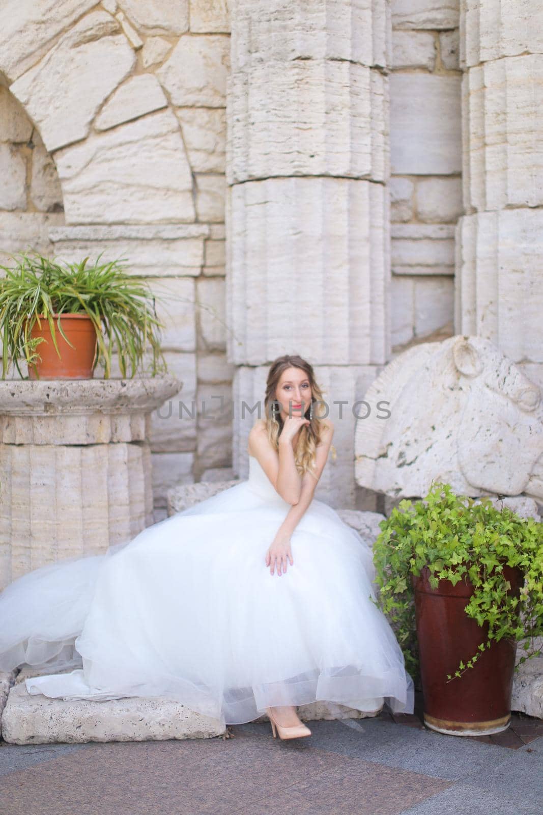 Caucasian charming fiancee sitting near ancient columns and wearing white dress. Concept of bridal photo session and wedding.