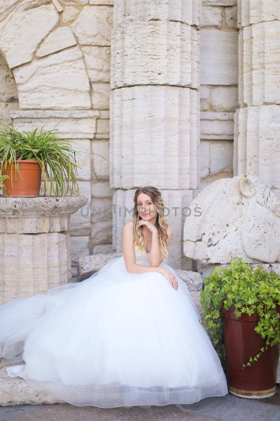Caucasian beautiful fiancee sitting near ancient columns and wearing white dress. Concept of bridal photo session and wedding.
