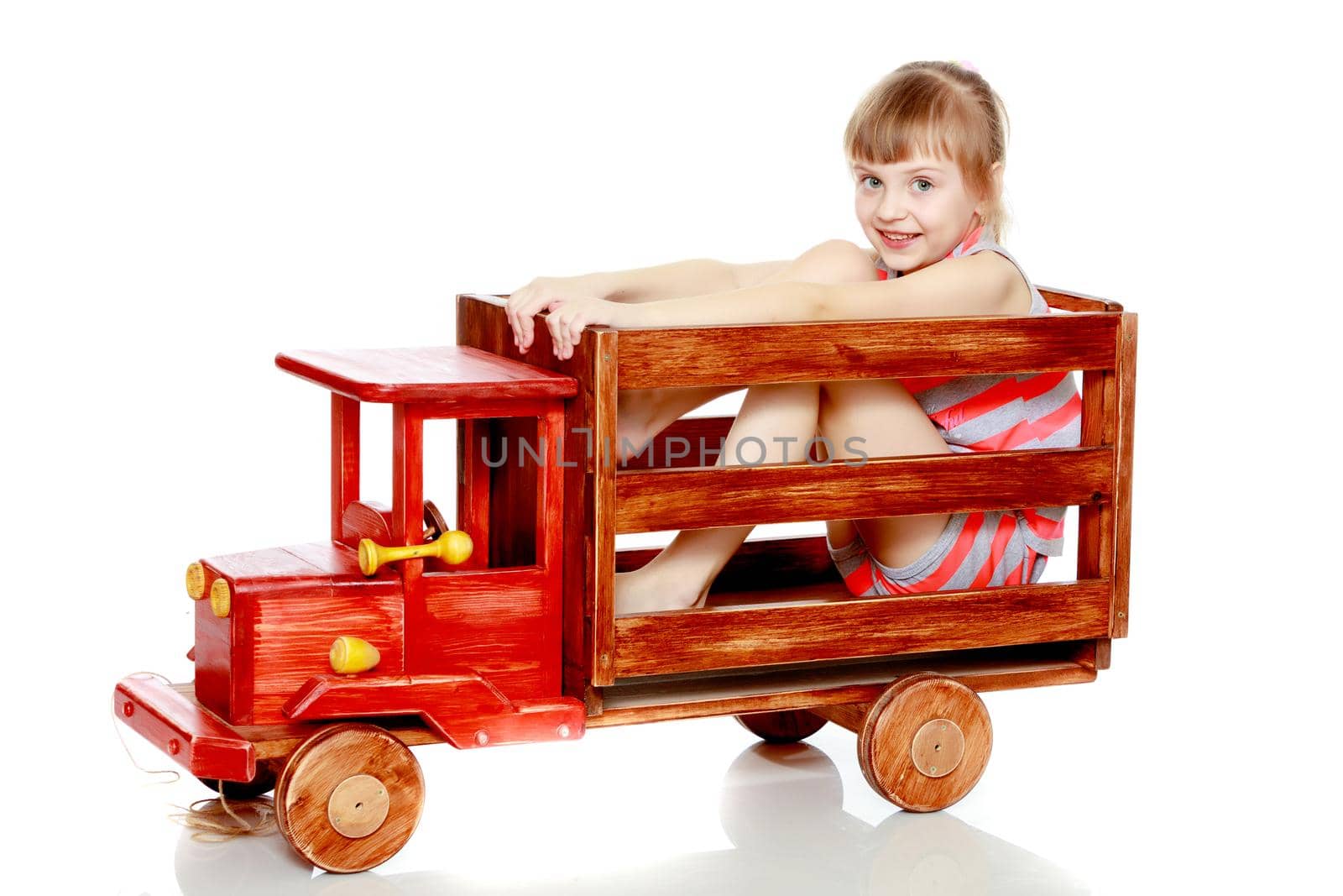Beautiful little caucasian girl with a short bangs and hair tucked into a tail. In striped in a T-shirt and shorts.The girl is sitting on a large toy wooden car.