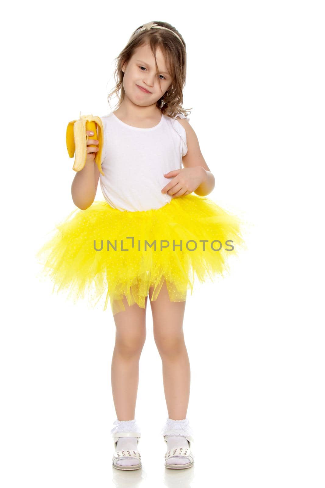 Joyful little blonde girl in a short yellow skirt and yellow bow on her head.She's holding a banana.Isolated on white background.