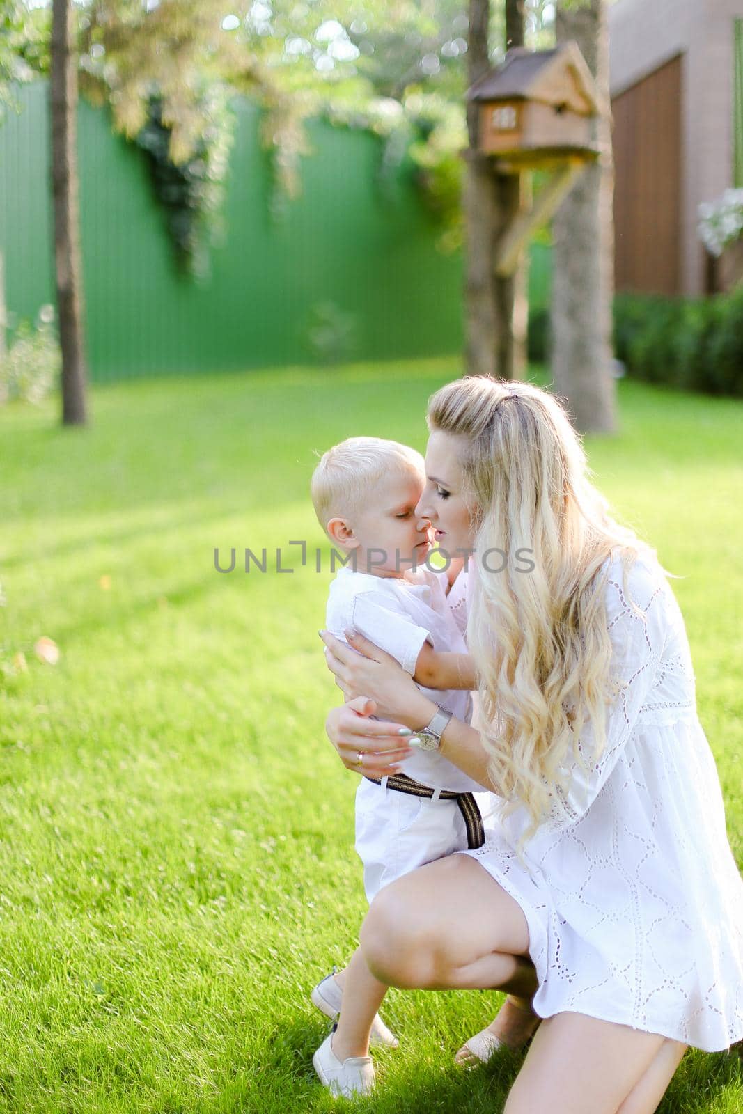 Young caucasian mother playing and sitting with little kid on grass in yard and kissing son. Concept of motherhood and child.
