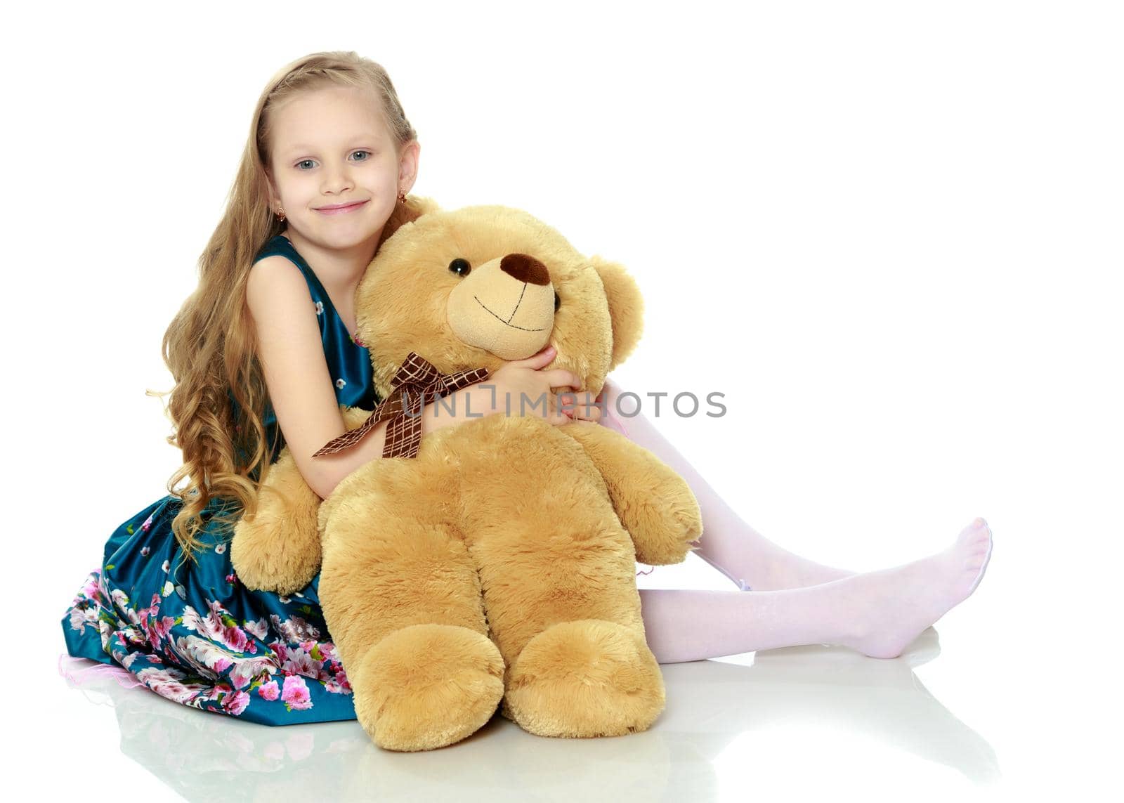 Beautiful little girl with long blond hair in a smart blue dress. The girl hugs a large teddy bear.
