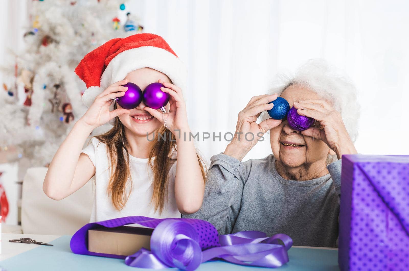 Smiling granddaughter in santa hat holding decorative balls like eyes with grandma at Christmas