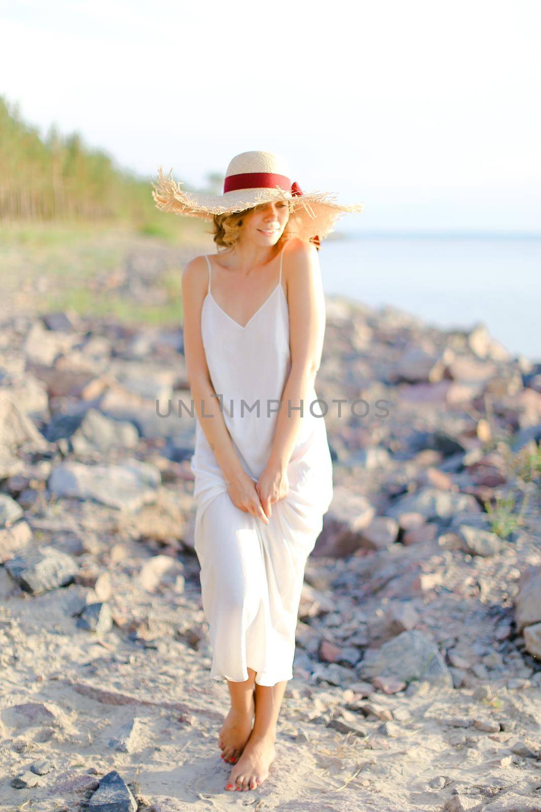 Young woman in white dress and hat with red ribbon standing on shingle beach. Concept of summer vacations and resort.