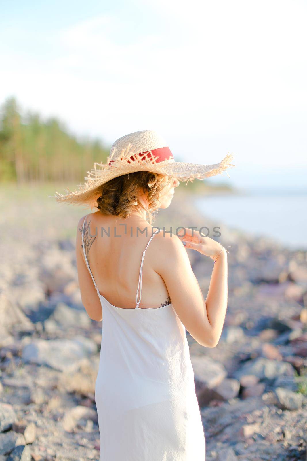 Back view of girl in hat with red ribbon walking on shingle beach. Concept of summer vacations and resort.