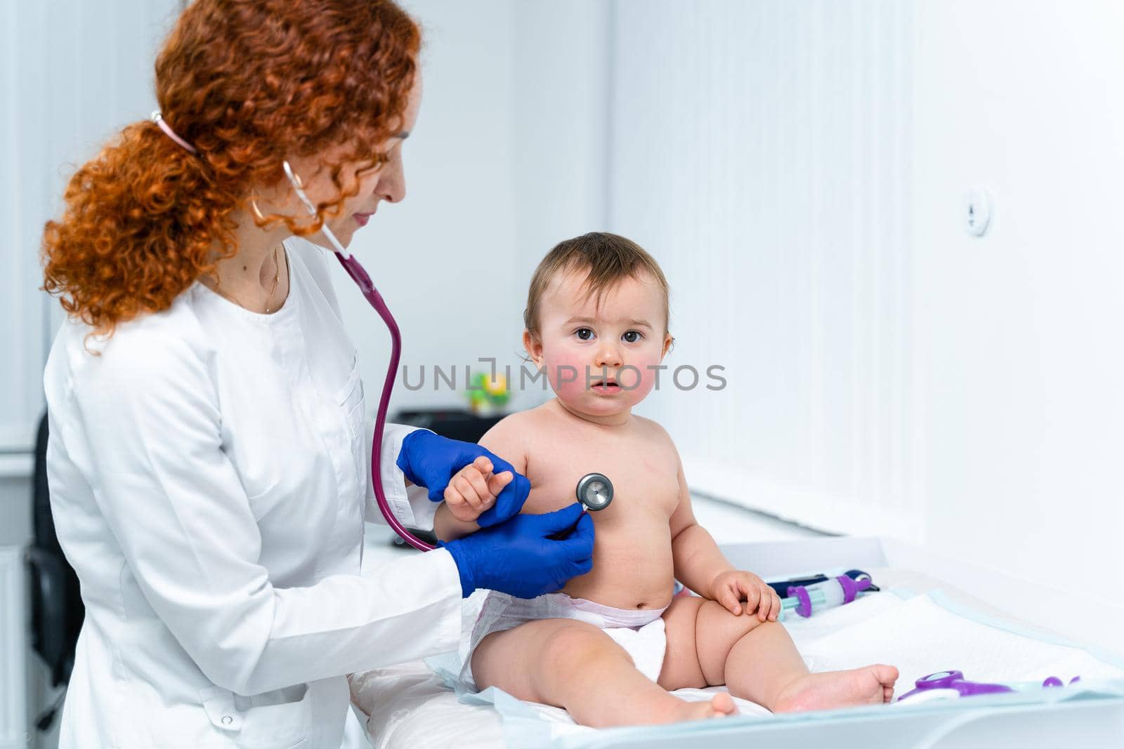 Red-haired female doctor examines little child for less than a year in a modern clinic. Pediatrician during examination baby in the hospital. Childrens healthcare. Diagnostics and treatment for kid.