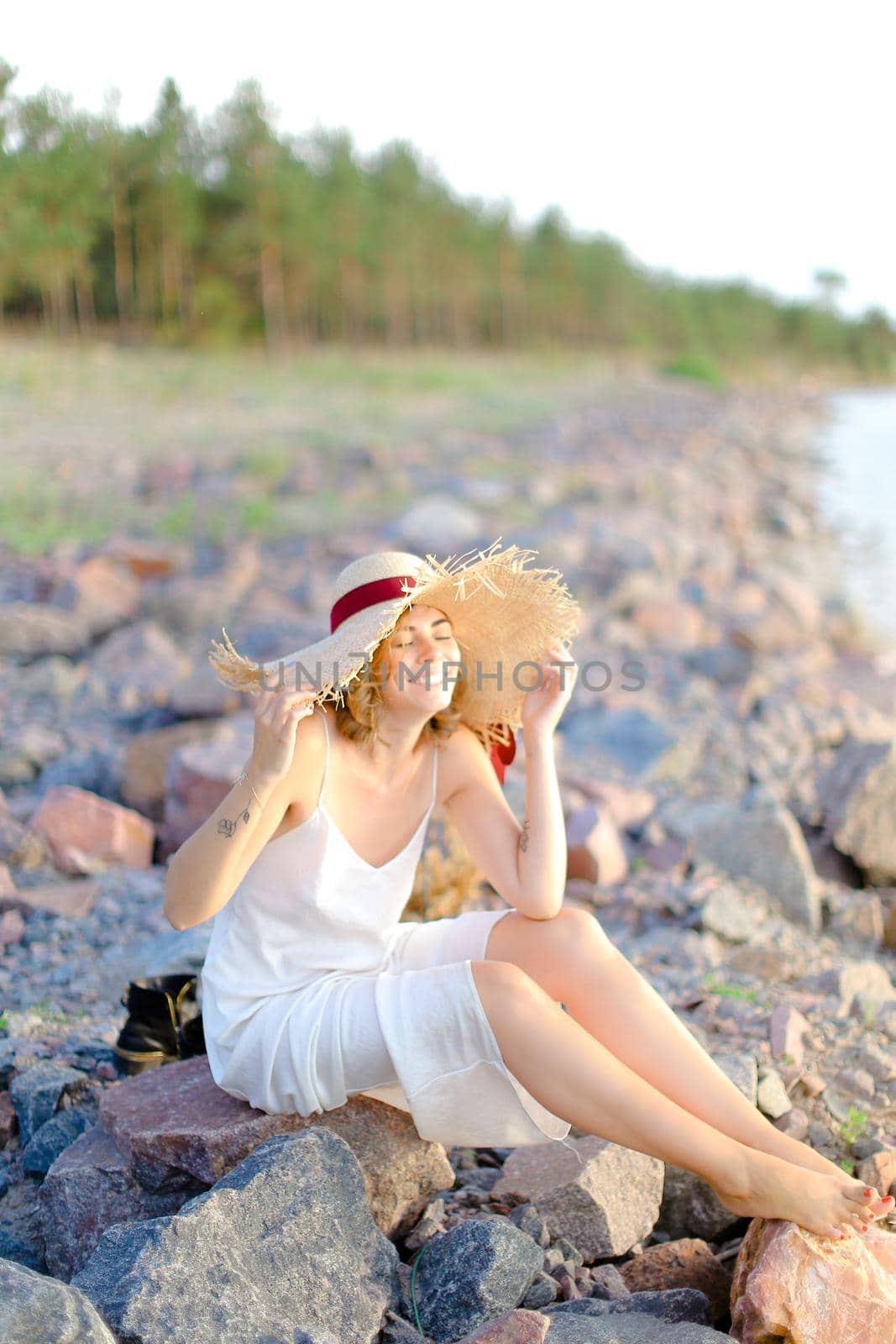 Caucasian pretty girl in hat sitting on shingle beach. Concept of summer vacations and beauty.