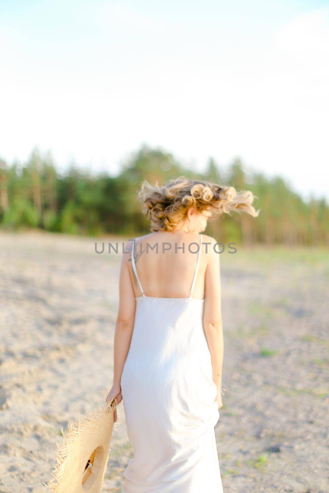 Back view of blonde girl walking on rocky beach with hant in hands and wearing dress. Concept of summer vacations and fashion.