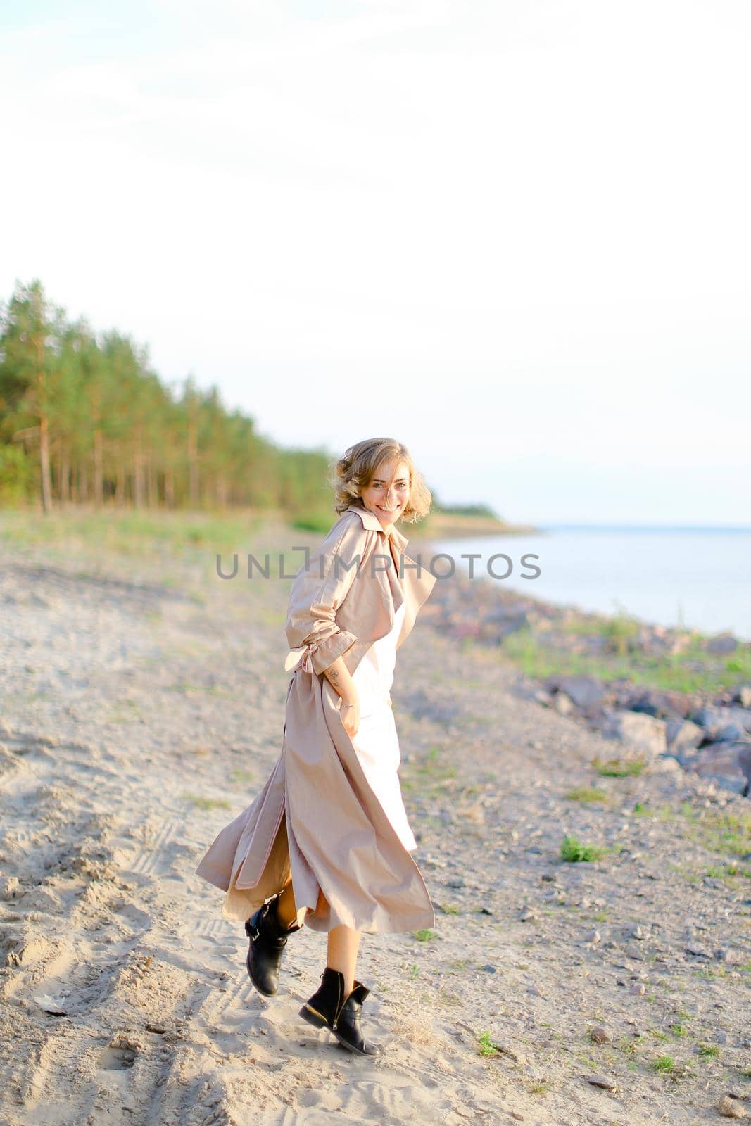 Back view of caucasian young female person walking on shingle beach and wearing summer coat. Concept of seasonal fashion and summer vacations.