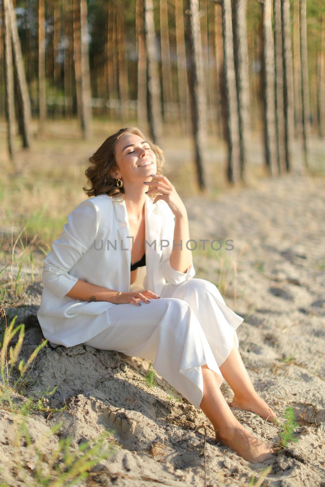 Young woman sitting on sand beach with trees in backgroundand wearing white clothes. Concept of freedom and summer vacations