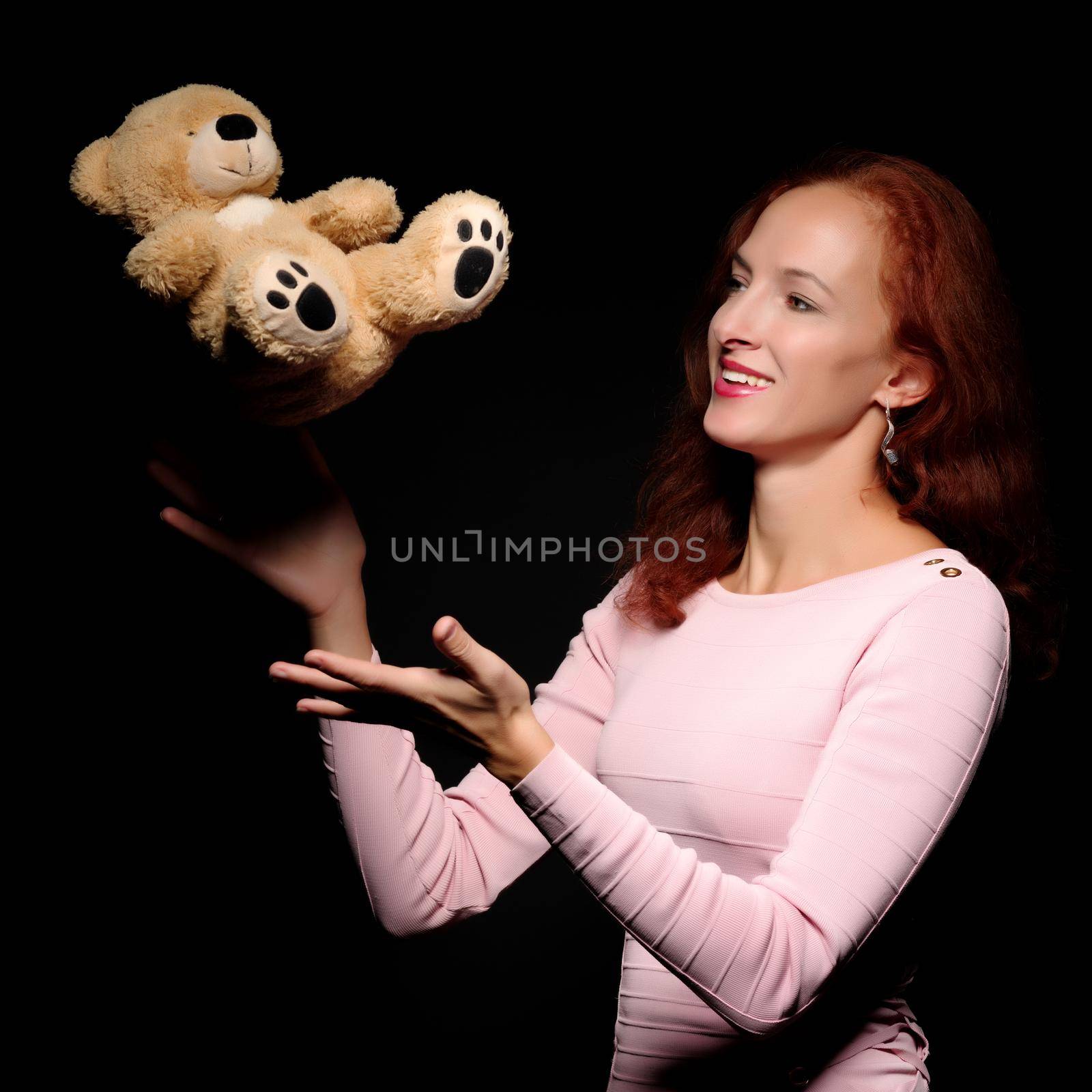 Fashion, artistic portrait of a beautiful redhead model girl, with long hair on a black background. Posing with a teddy bear.