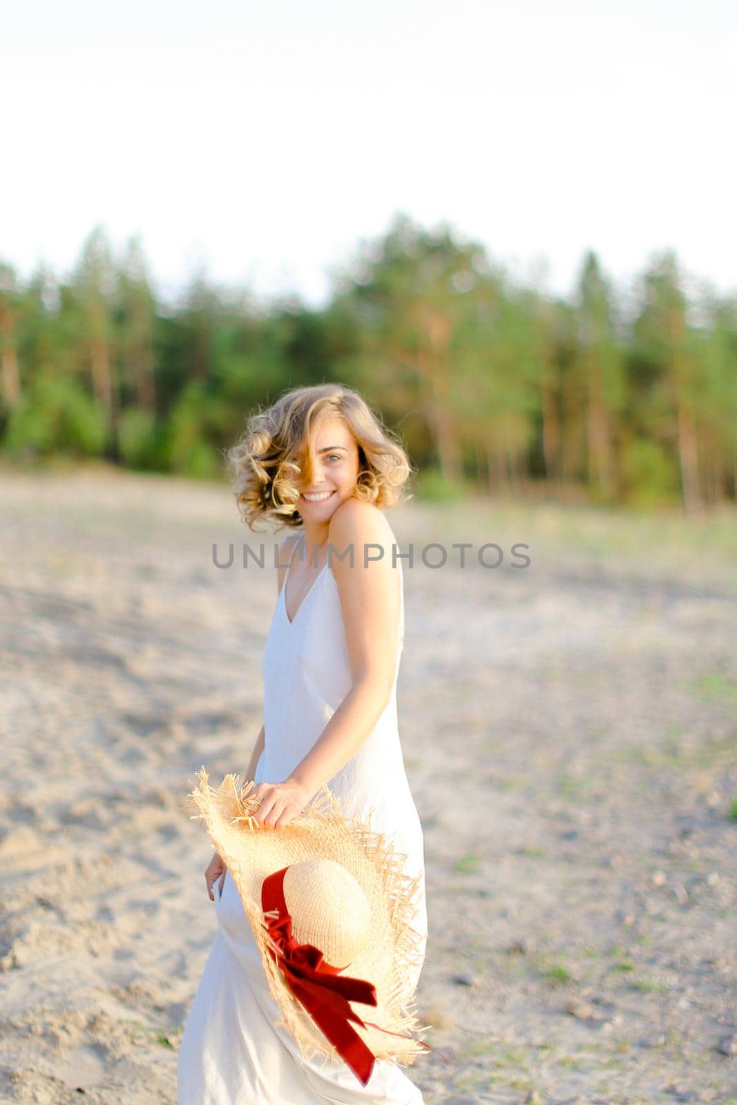 American blonde girl walking on rocky beach with hant in hands and wearing dress. Concept of summer vacations and fashion.