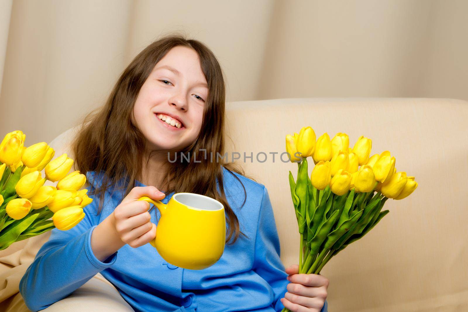 A charming school girl is sitting on the couch with a mug in her hands. Concept of happy people, healthy eating.