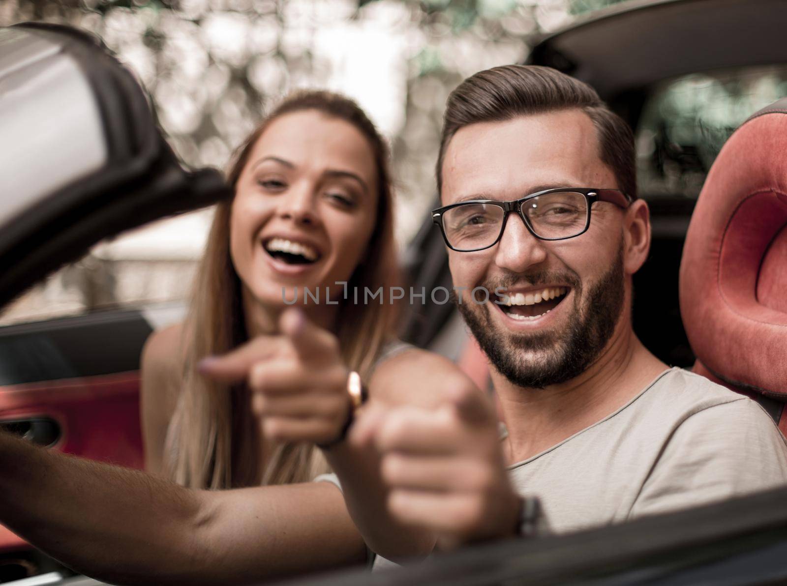 happy couple sitting inside a convertible and pointing at you by asdf