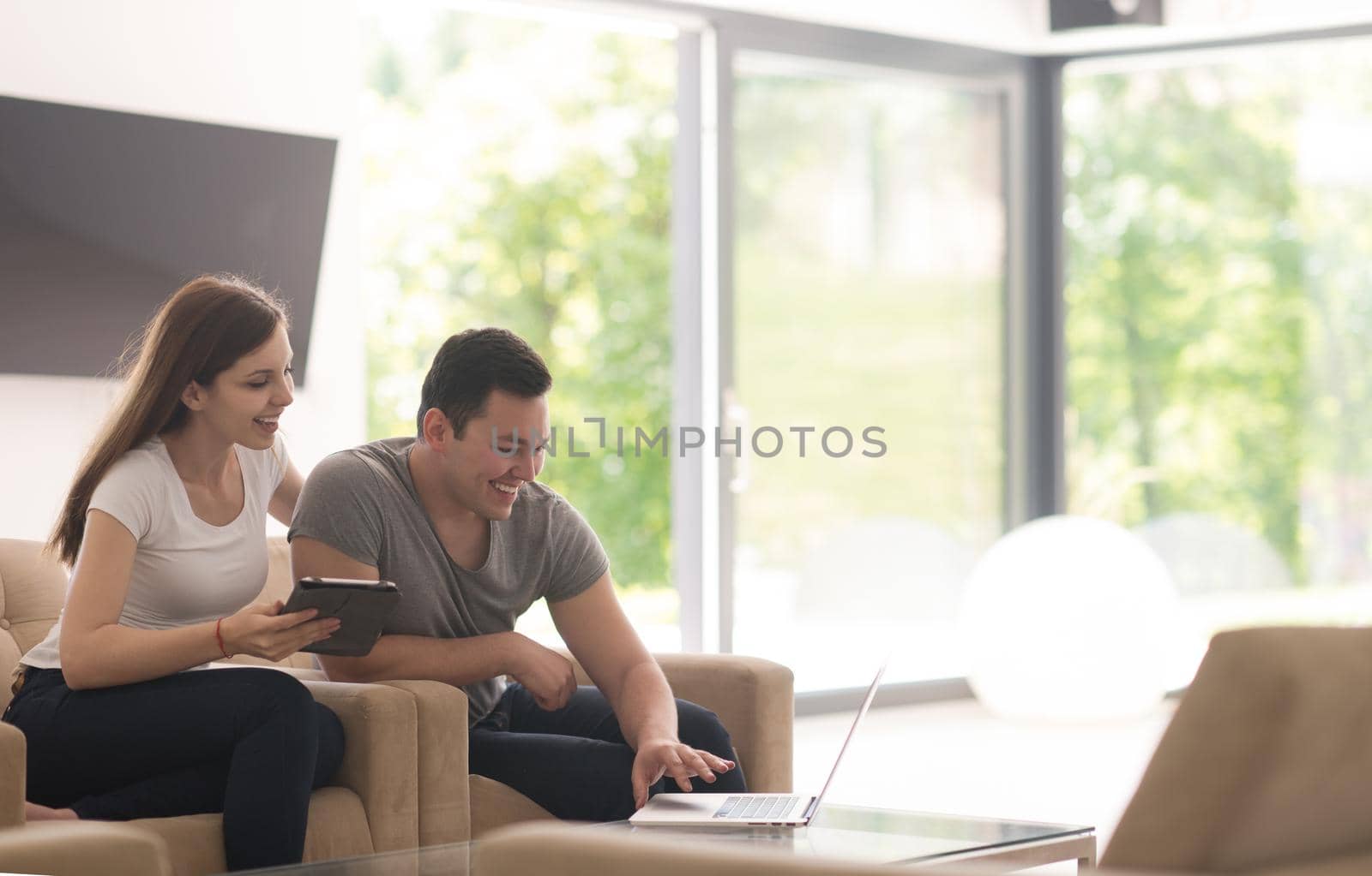 Young couple relaxing at luxurious home with tablet and laptop computers reading in the living room on the sofa couch.