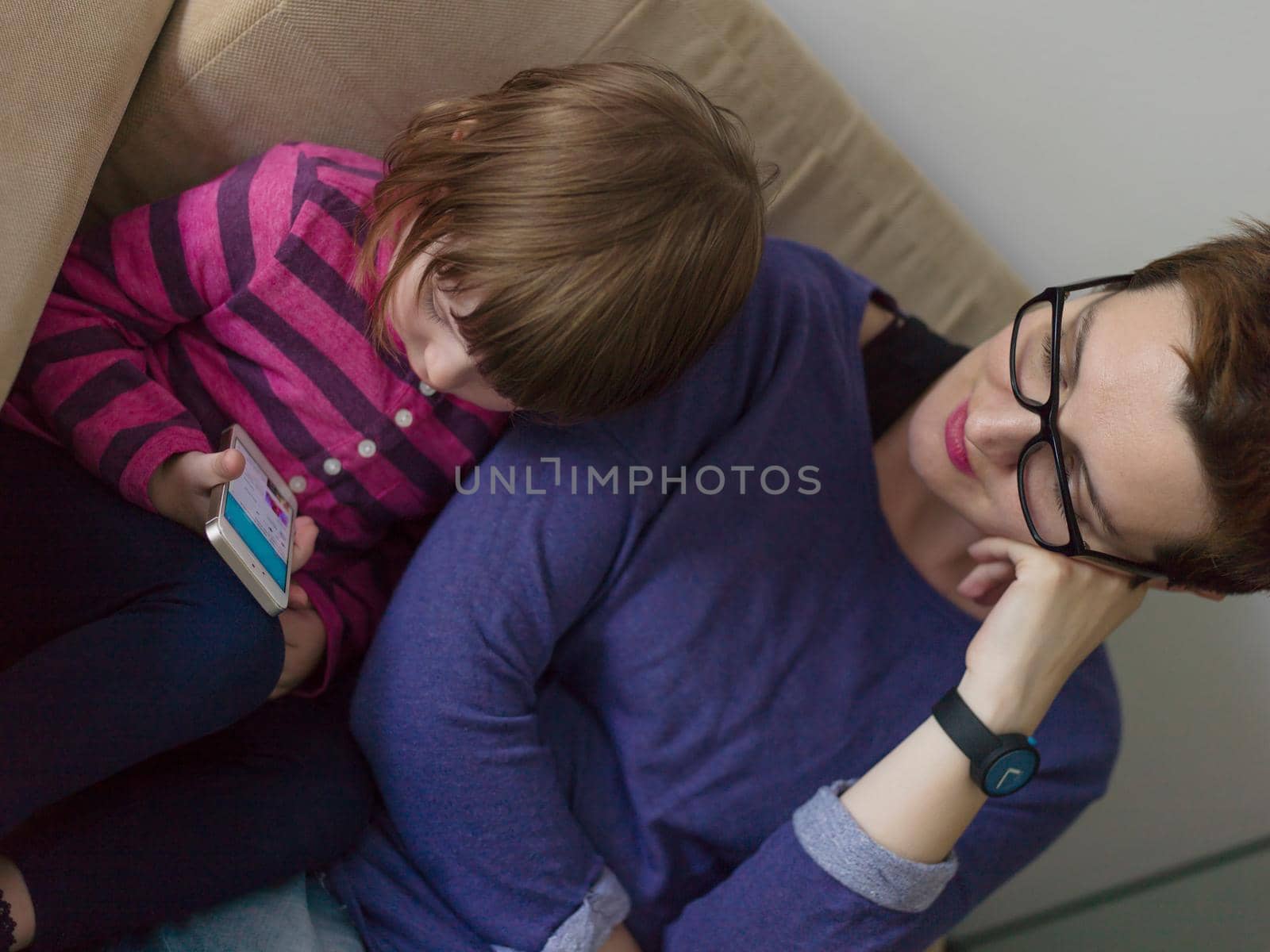 Beautiful young mother and her cute little daughter are using a mobile phone and smiling, sitting on sofa at home