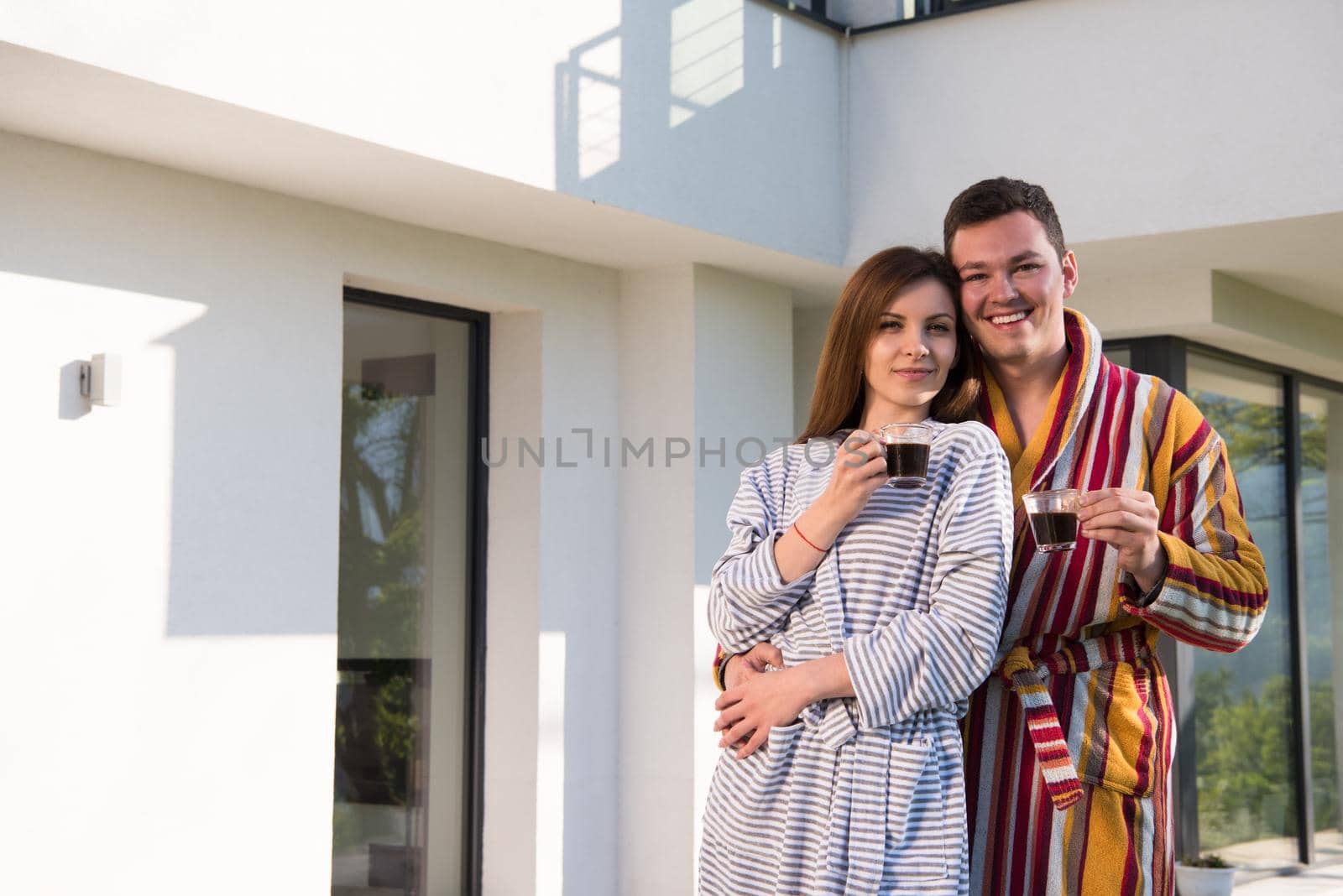 Young beautiful couple in bathrobes are enjoying morning coffee in front of their luxury home villa