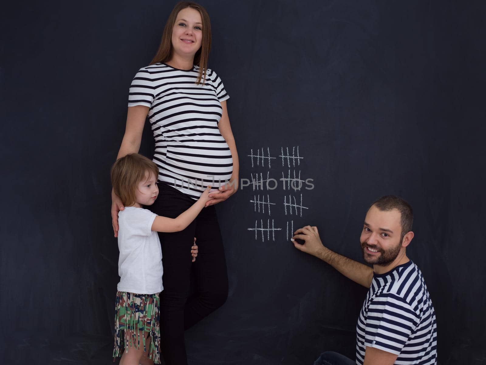 young pregnant couple with cute little daughter accounts week of pregnancy and writing them with chalk on blackboard