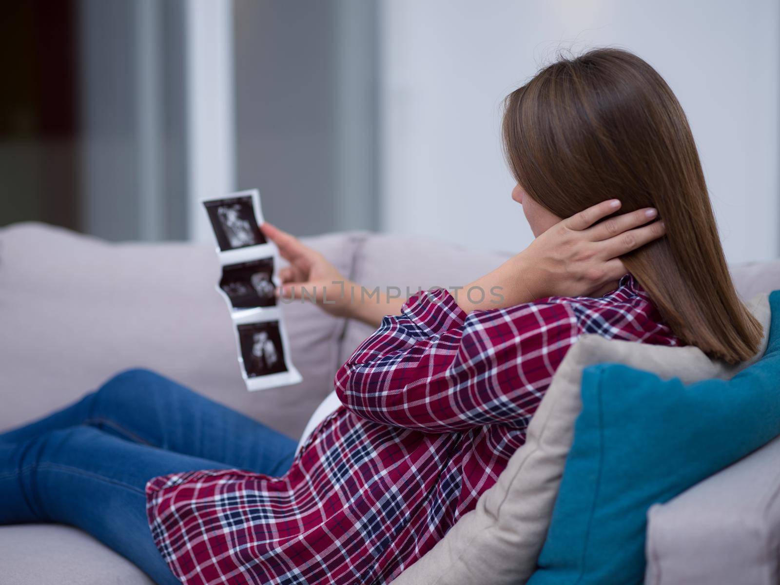 Young pregnant woman looking baby's ultrasound photo while relaxing on sofa at home