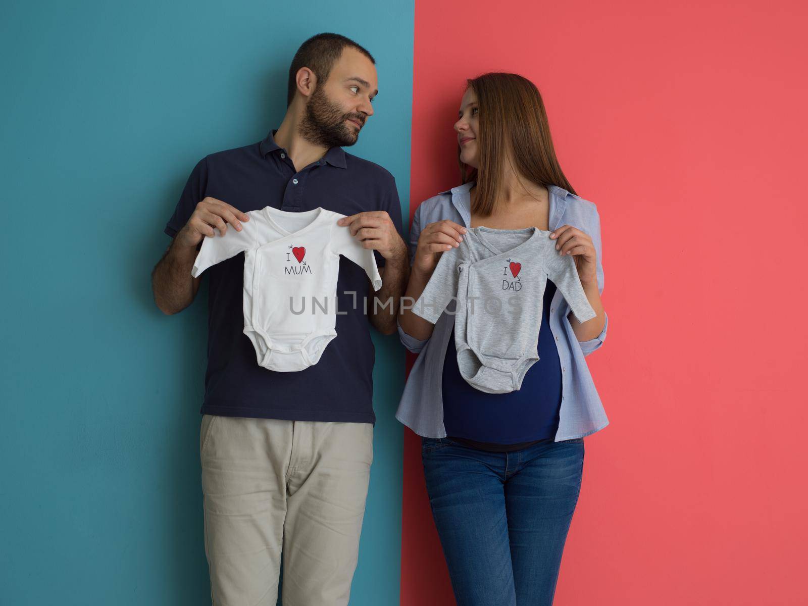 Beautiful pregnant woman and her husband expecting baby holding baby bodysuits and smiling over colorful background