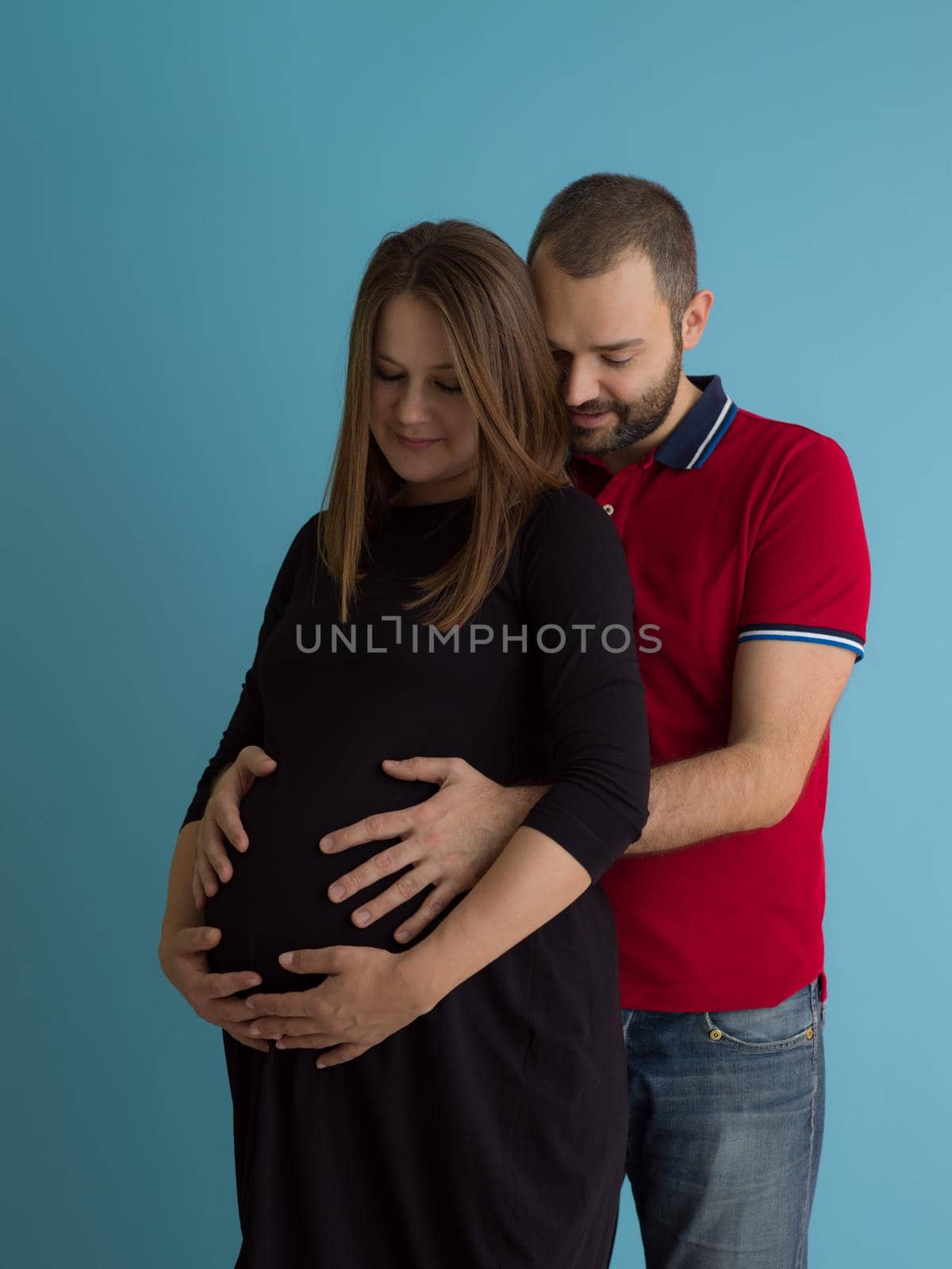 Portrait of a happy young couple,man holding his pregnant wife belly isolated over blue background