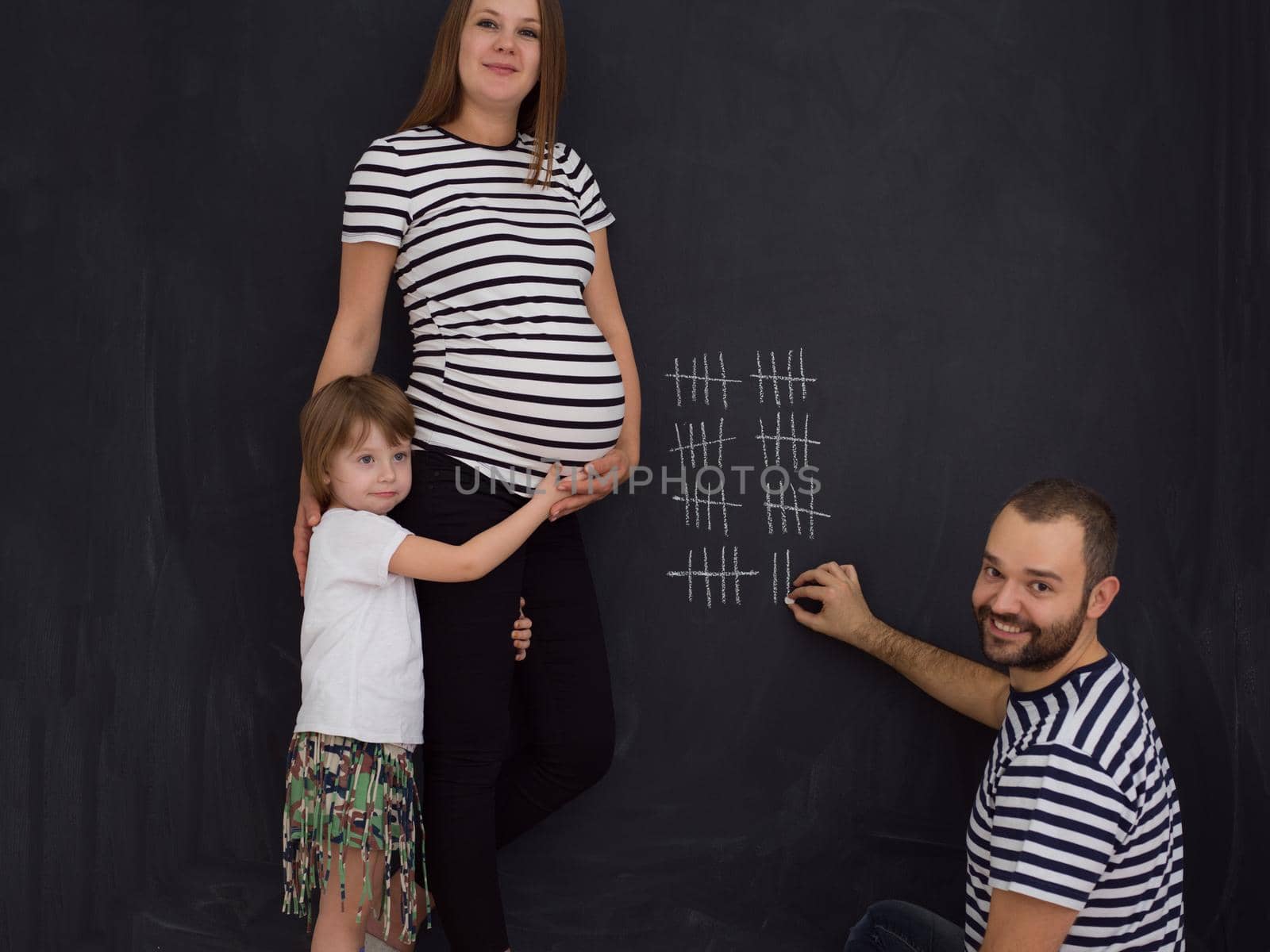 young pregnant couple with cute little daughter accounts week of pregnancy and writing them with chalk on blackboard