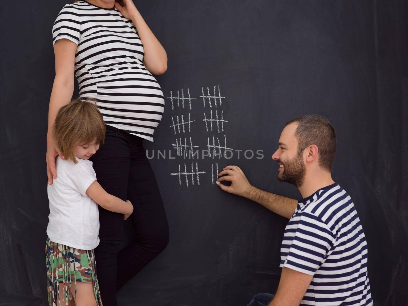 young pregnant couple with cute little daughter accounts week of pregnancy and writing them with chalk on blackboard