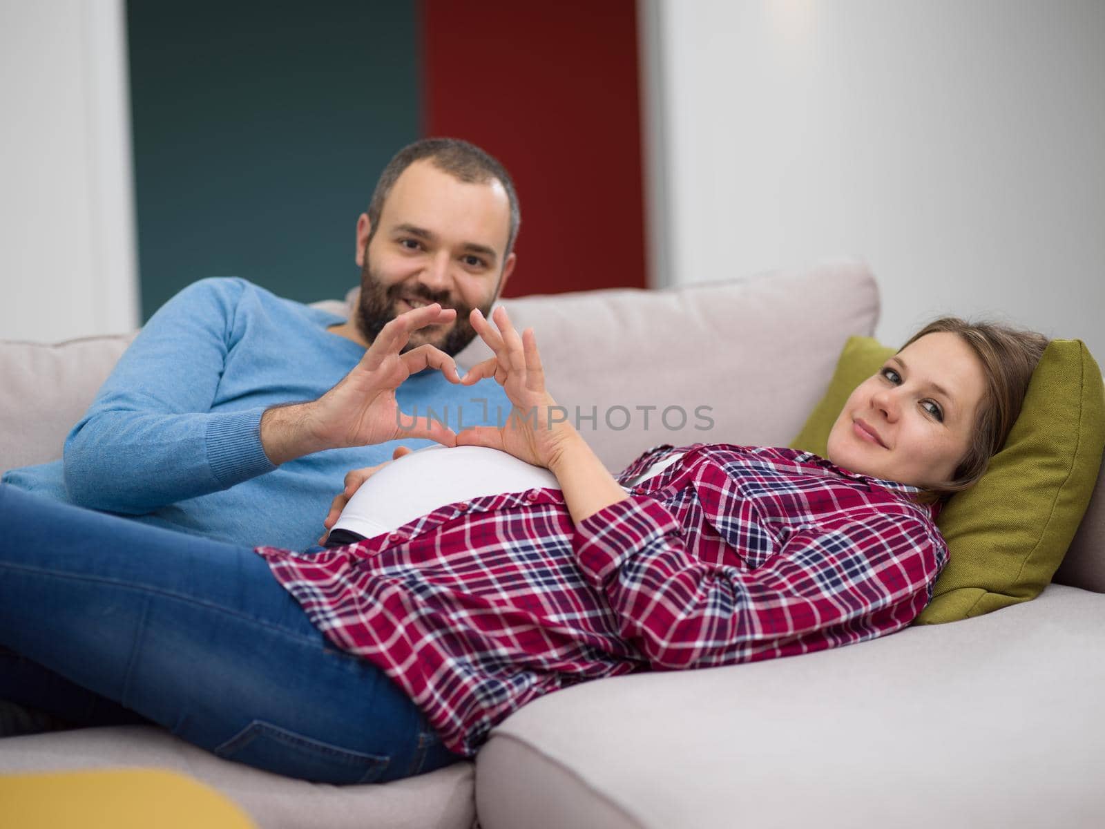 Happy man and pregnant woman showing heart sign with fingers while relaxing on the sofa at home