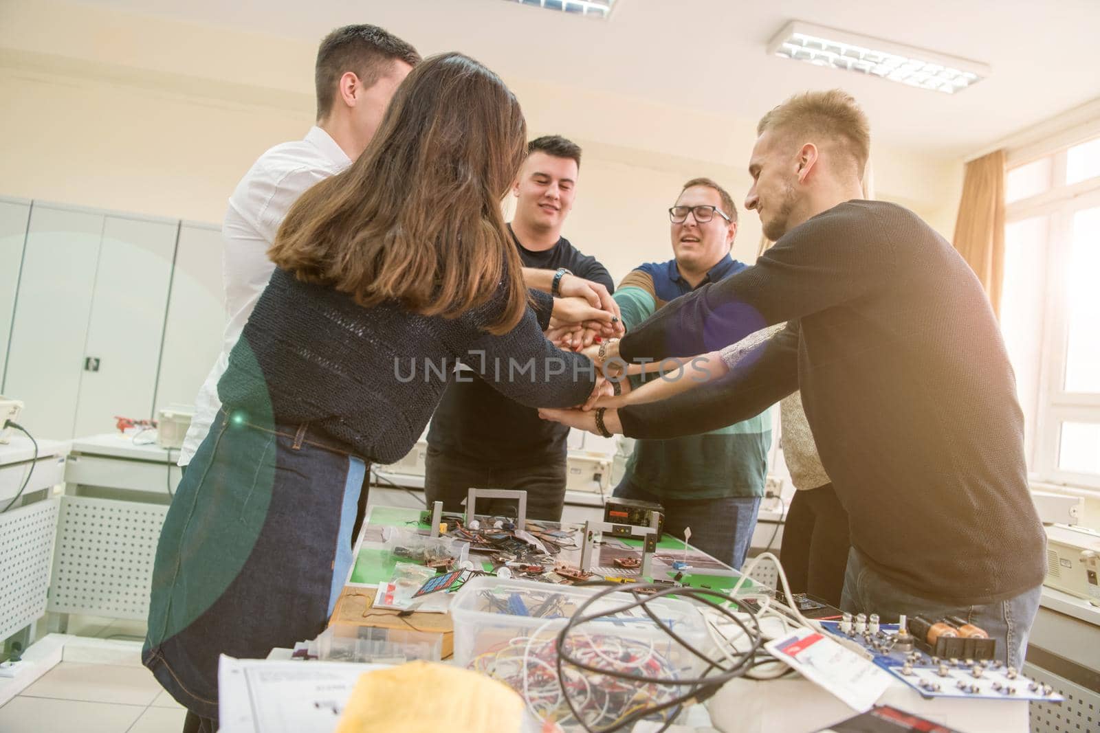 Group of young students in electronics classroom celebrating successfully finished project with holding their hands together, education and technology concept