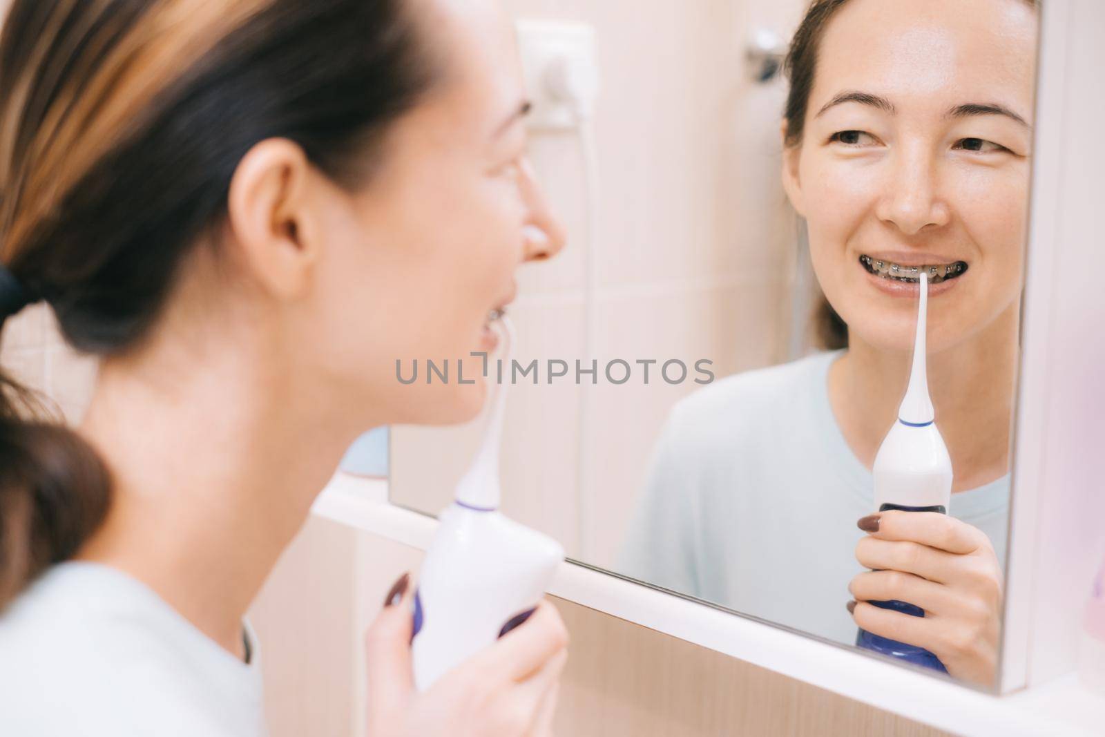 Young woman with braces on her teeth brushing her teeth with by using a irrigate, before mirror. Dental and oral care.