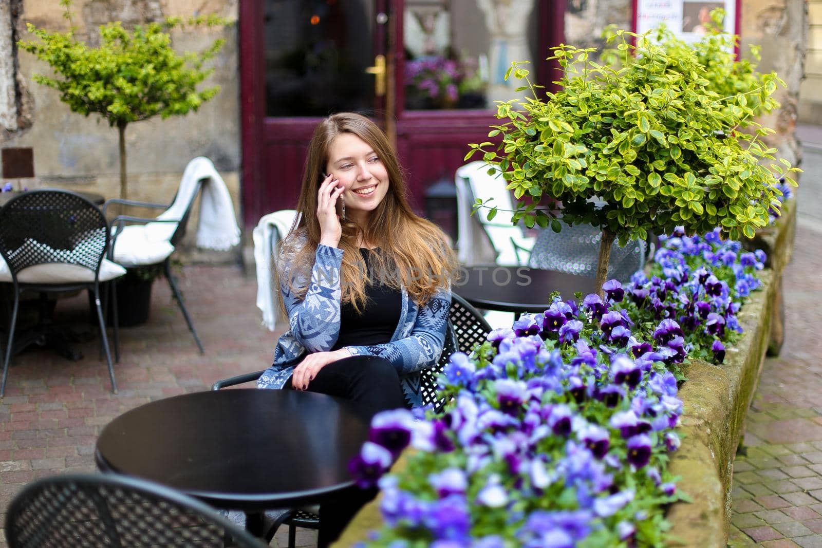 Young woman speaking by smartphone and sitting at street cafe near blue flowers and green plants. Concept of cozy place in open air and resting.