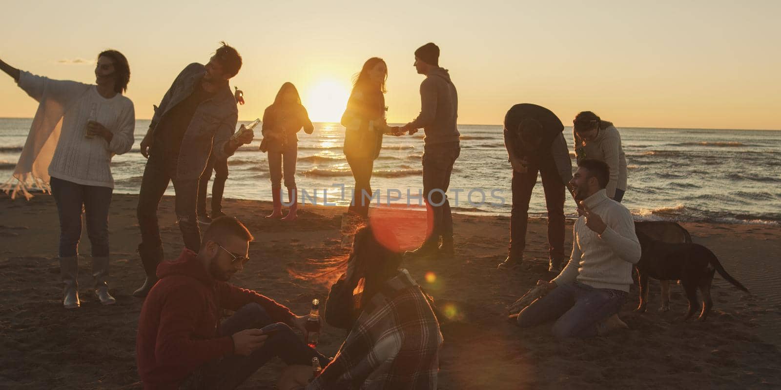 Happy Carefree Young Friends Having Fun And Drinking Beer By Bonefire On The Beach As The Sun Begins To Set