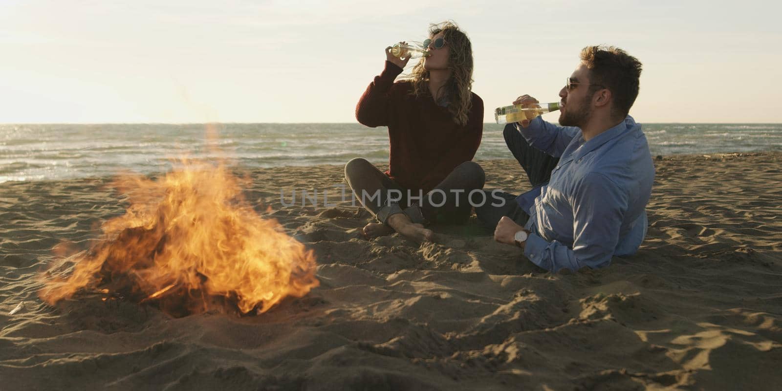 Loving Young Couple Sitting On The Beach beside Campfire drinking beer by dotshock