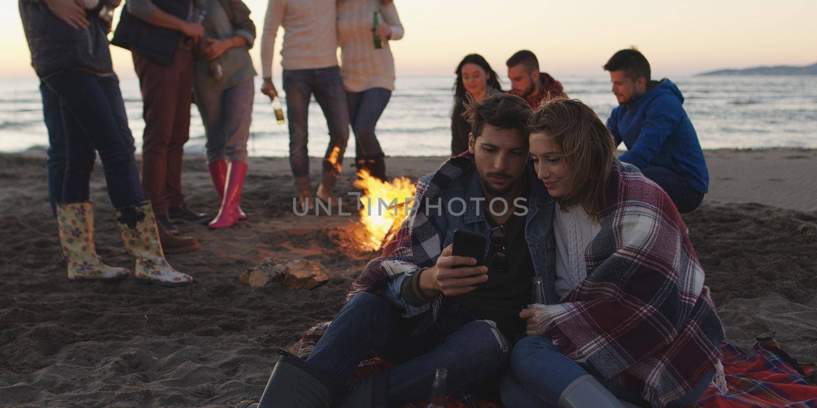 Couple using cell phone during beach party with friends drinking beer and having fun