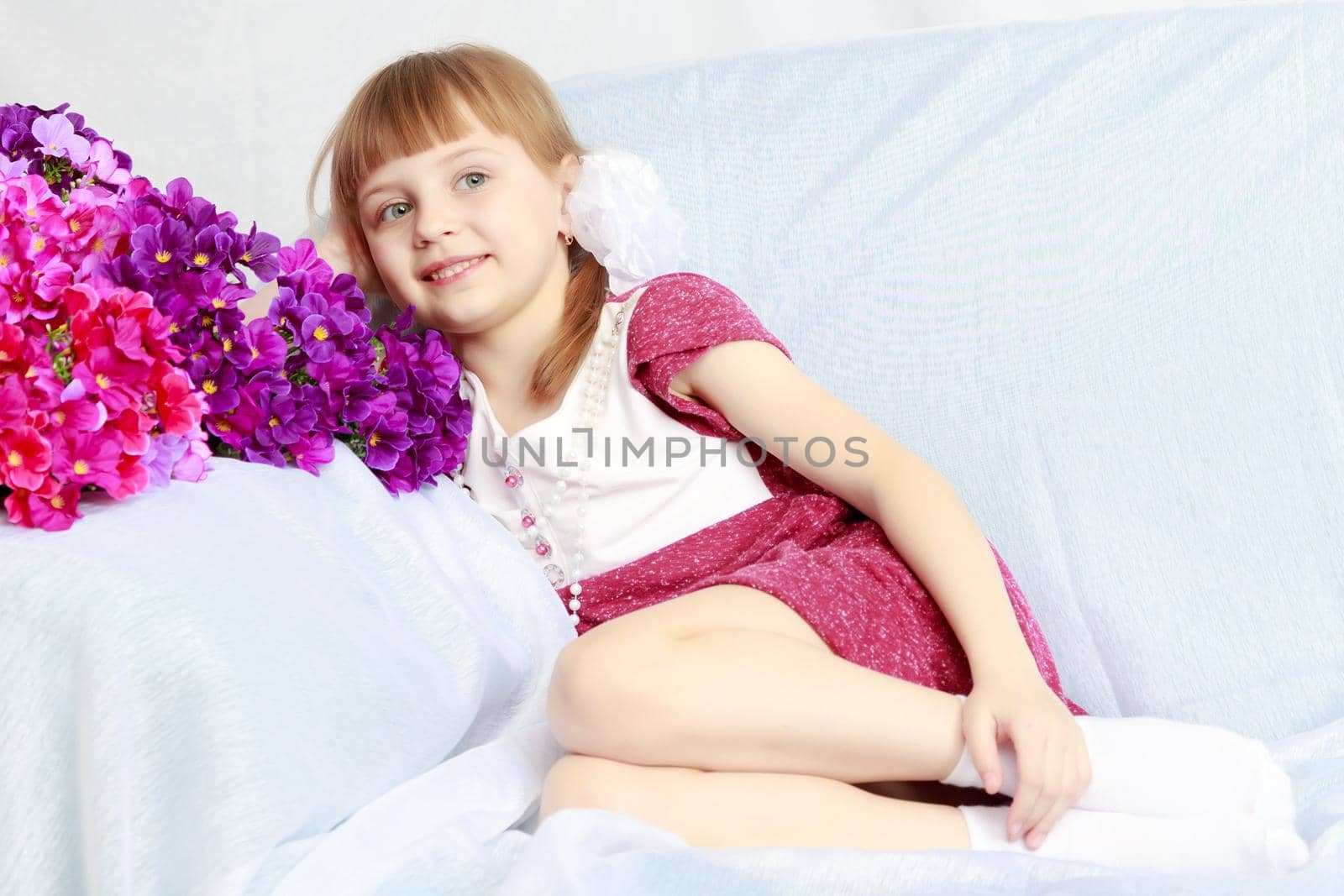 Beautiful little blonde girl with short bangs and pigtails on her head in a good mood.She sits next to a bouquet of flowers.