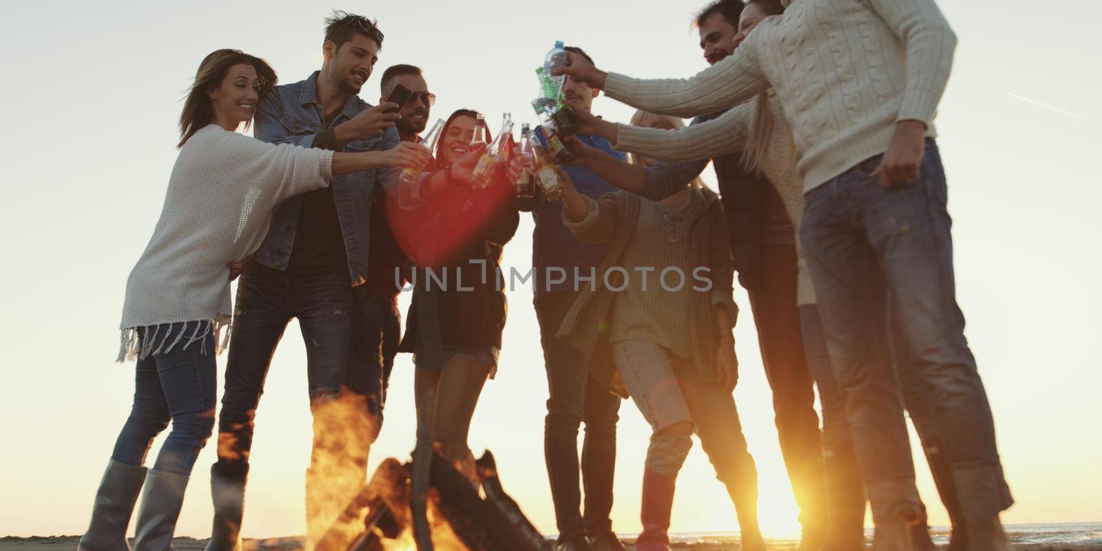 Young Friends Making A Toast With Beer and having fun Around Campfire at beach party on autumn day