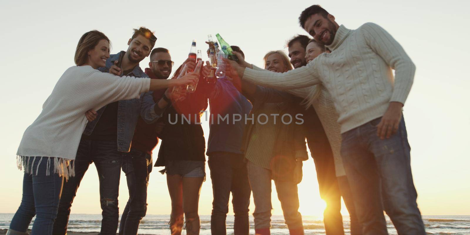 Young Friends Making A Toast With Beer and having fun Around Campfire at beach party on autumn day